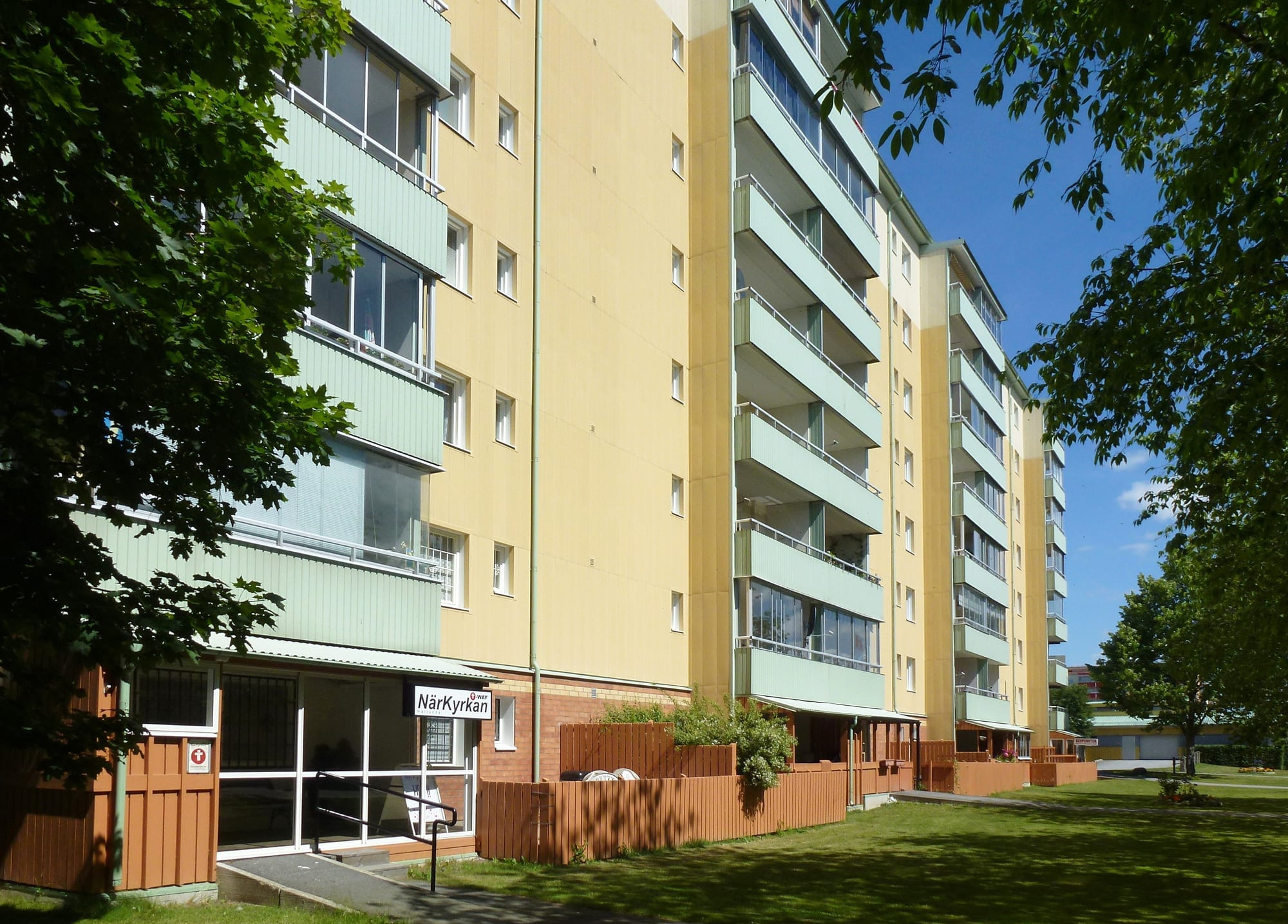 A sunlit multi-storey apartment block with balconies, painted in yellow and pale green, surrounded by trees and lawn