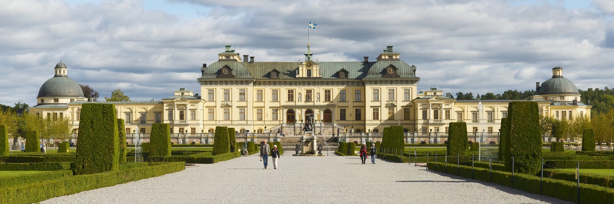 A grand yellow baroque palace with green domed towers behind formal gardens