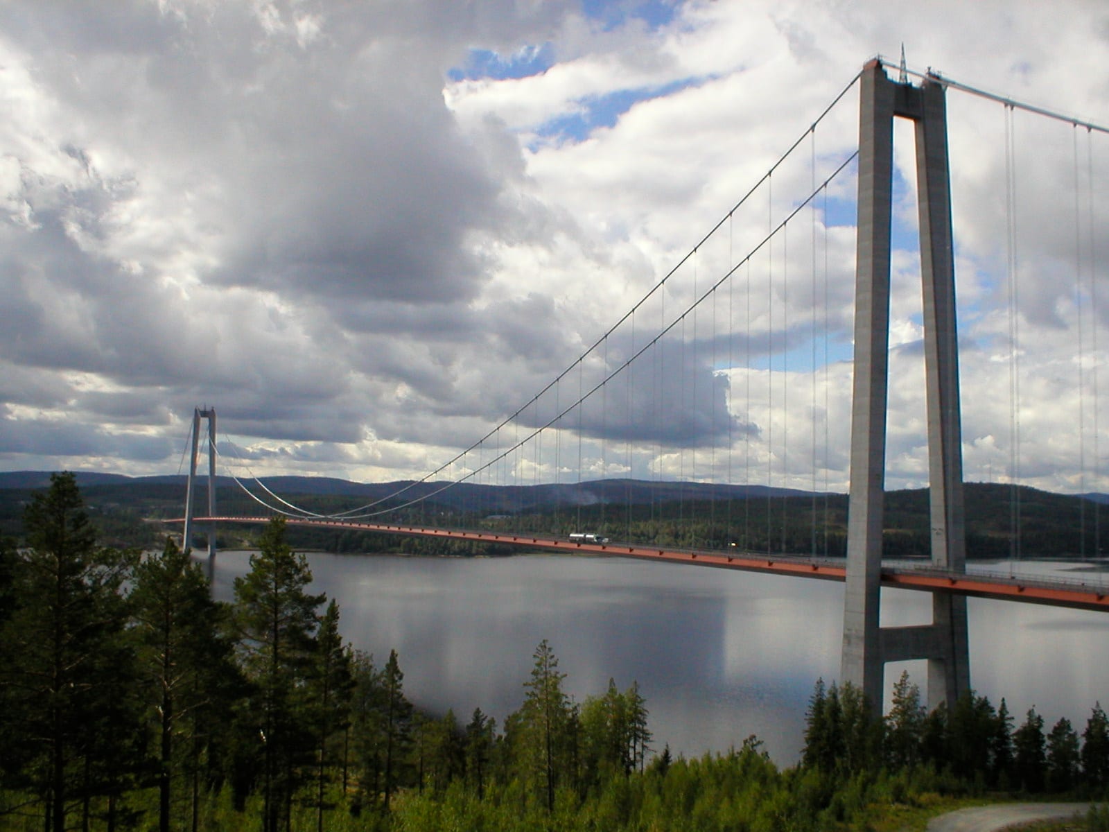 A tall suspension bridge spanning a wide body of water between forested hills