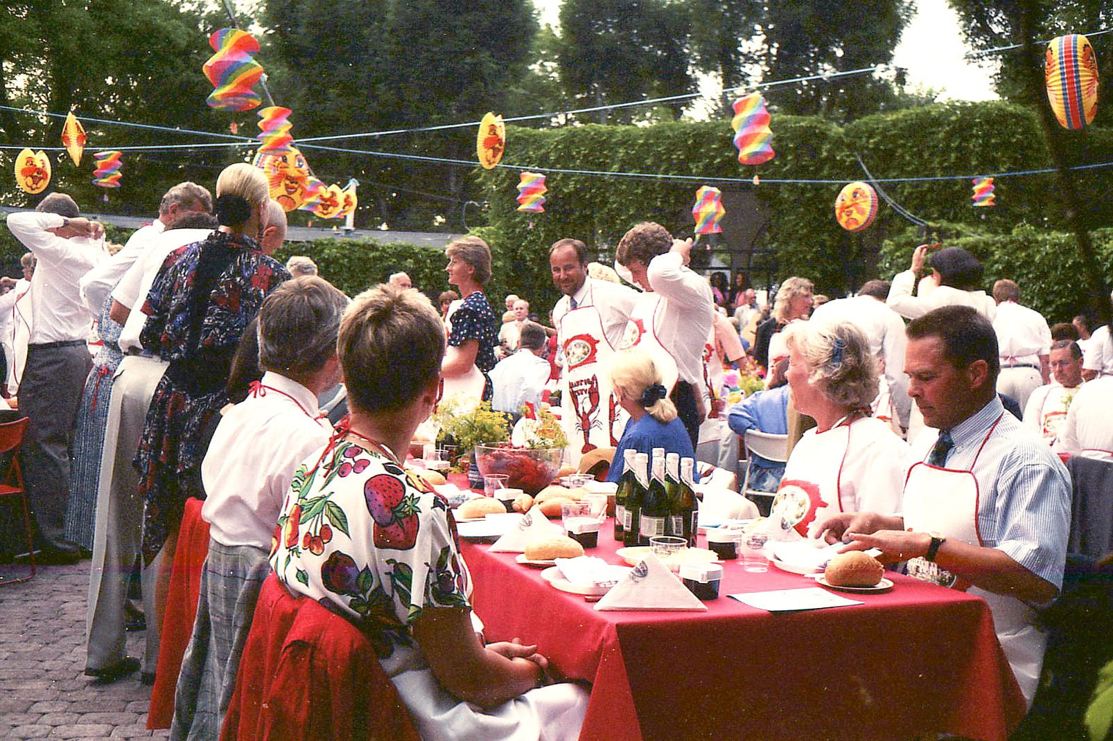Guests in paper bibs sit at long tables with red cloths, bread and bottles, under strings of colourful paper lanterns in a garden