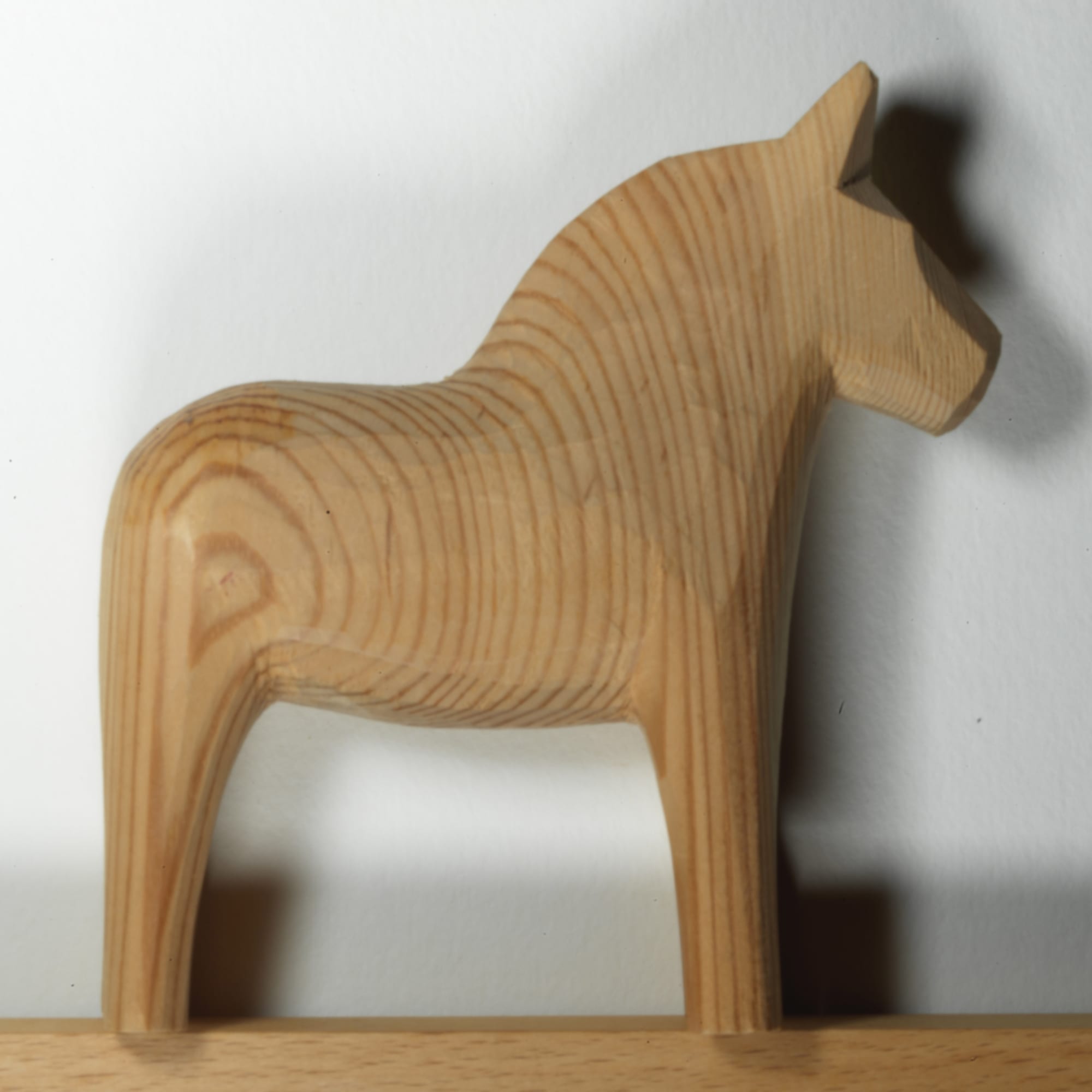 A plain, unpainted wooden horse carved from pale pine with visible grain, standing on a wooden shelf against a white wall