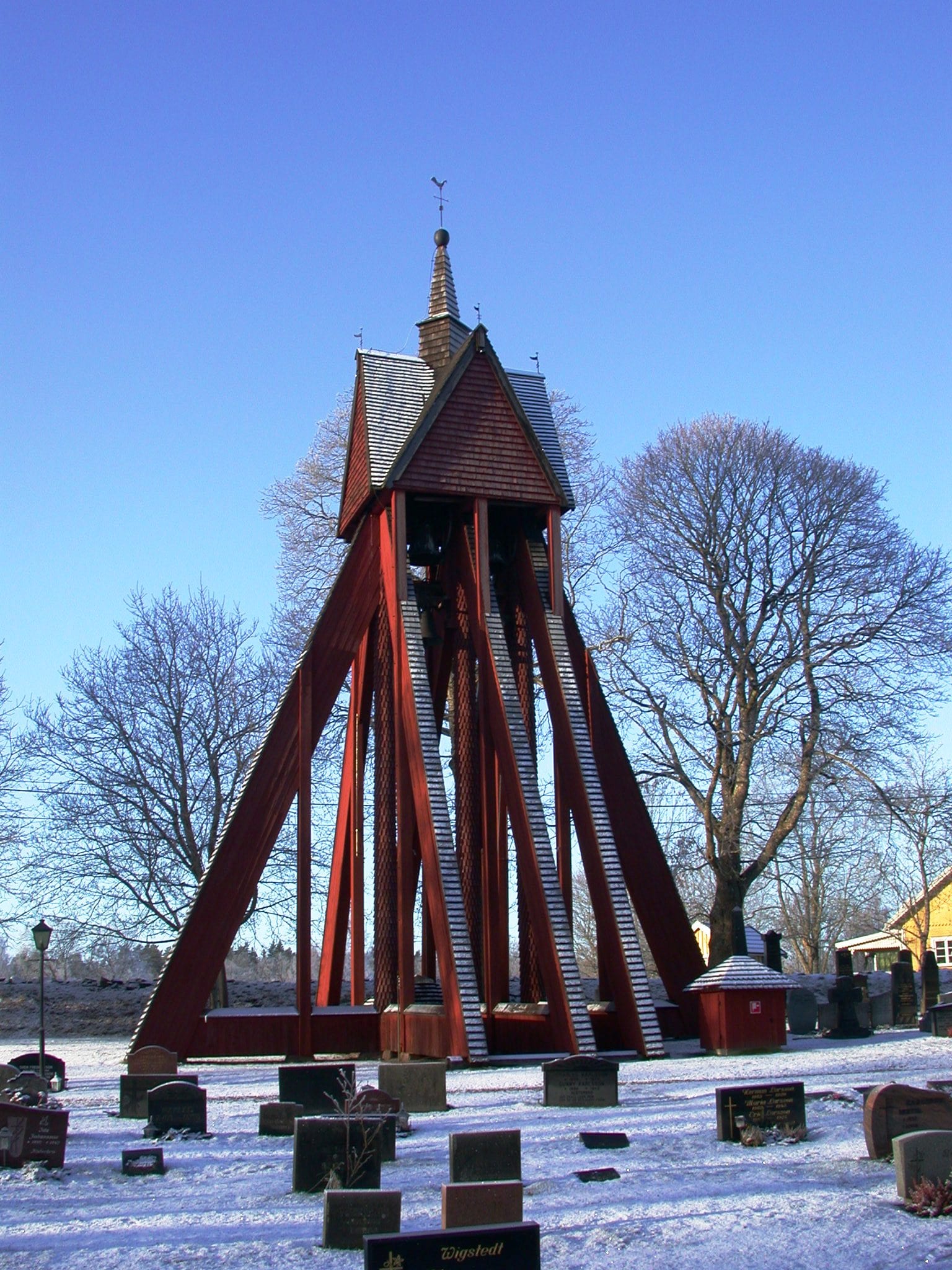 A tall red wooden bell tower with steeply angled timber supports and a shingled roof stands in a snowy churchyard with gravestones in front and bare trees behind, under a clear blue sky