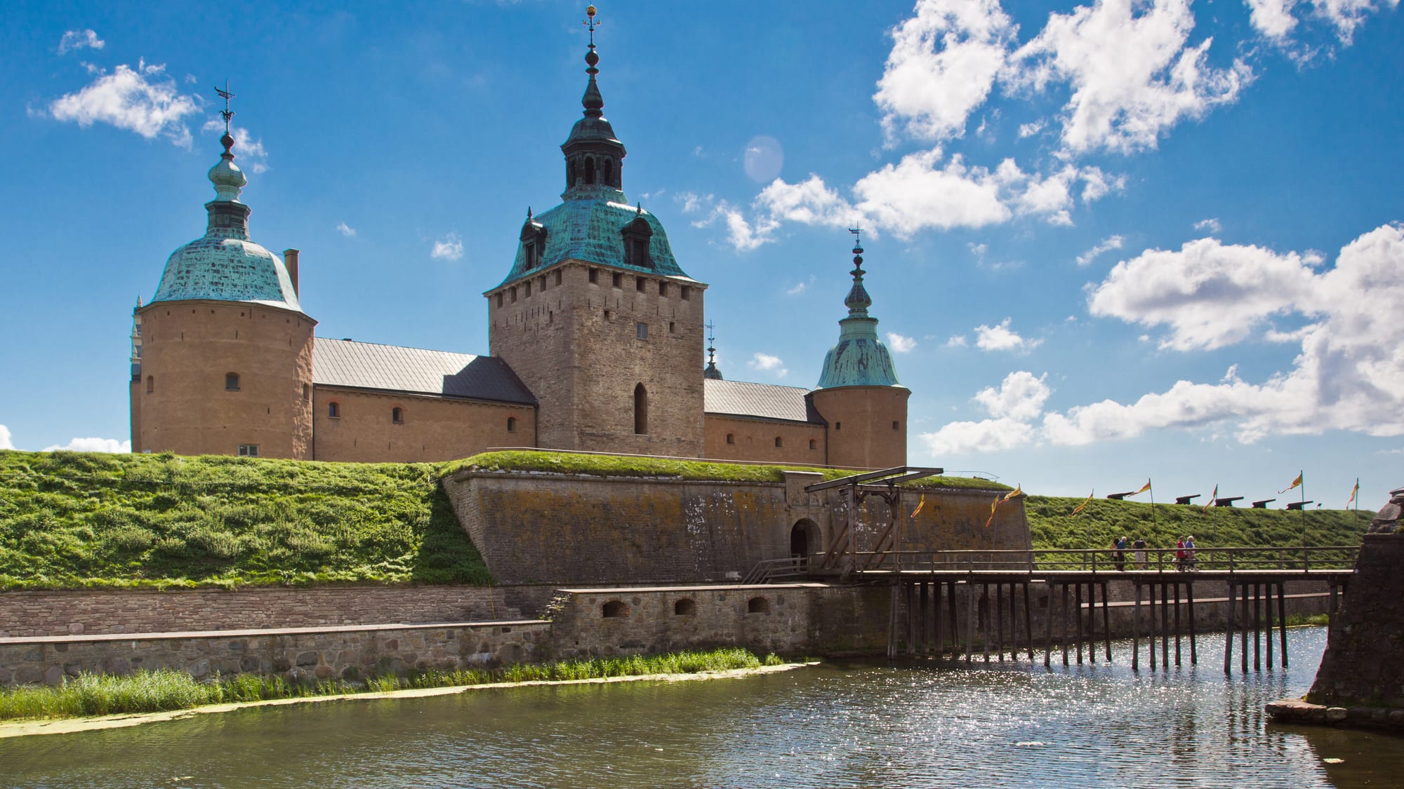 A stone castle with green copper-domed round towers standing above grassy ramparts, a moat and a wooden drawbridge under a blue sky