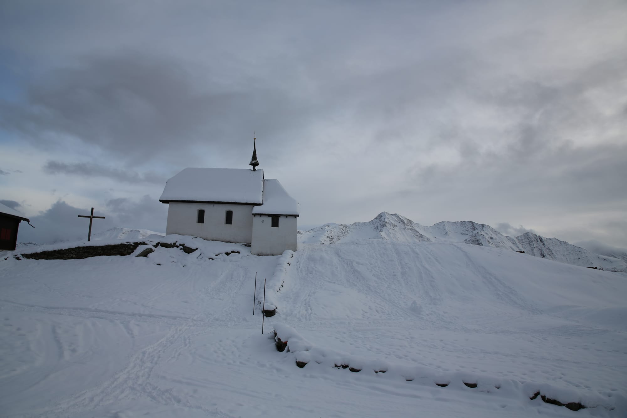 A small white chapel with a slim spire stands on a snow-covered rise beside a wooden cross, with snowy Alpine ridges behind under a cloudy sky