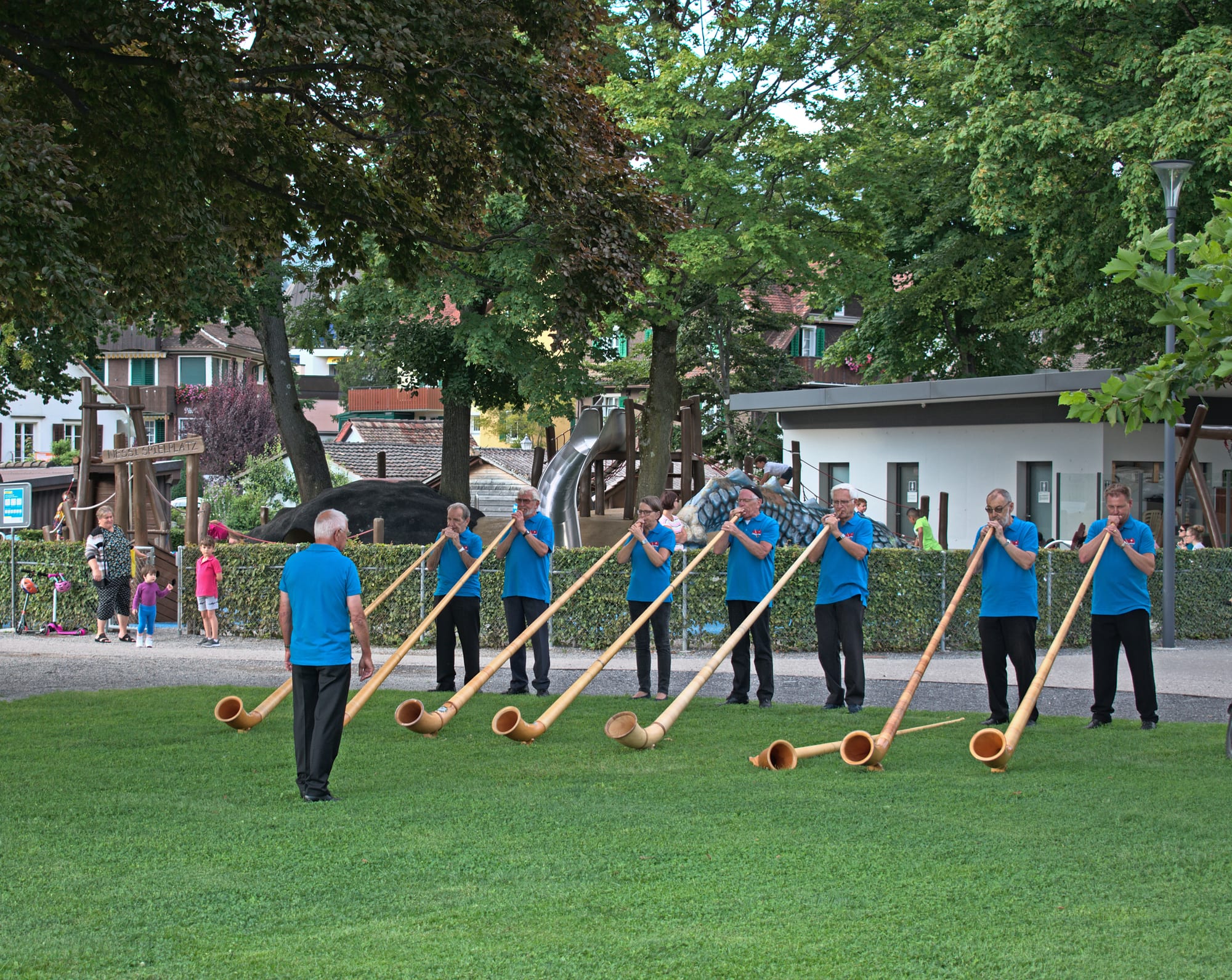 A row of men and women in matching blue shirts play long wooden alphorns resting on a lawn, while a conductor stands facing them, with trees and buildings behind