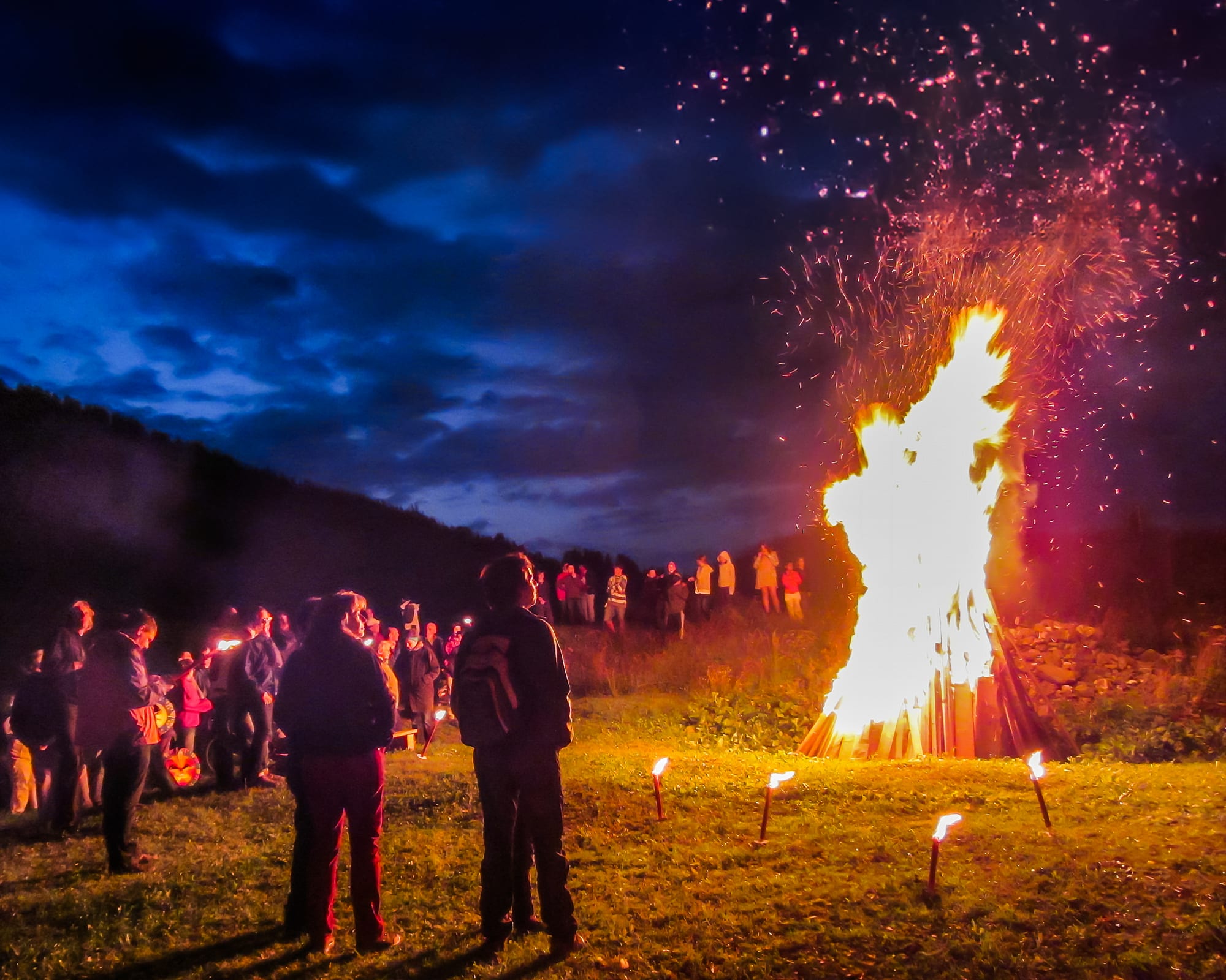 A large bonfire throws sparks into a deep blue night sky on a grassy hillside, with small torches in the grass and a crowd of people, seen mostly from behind, standing and watching