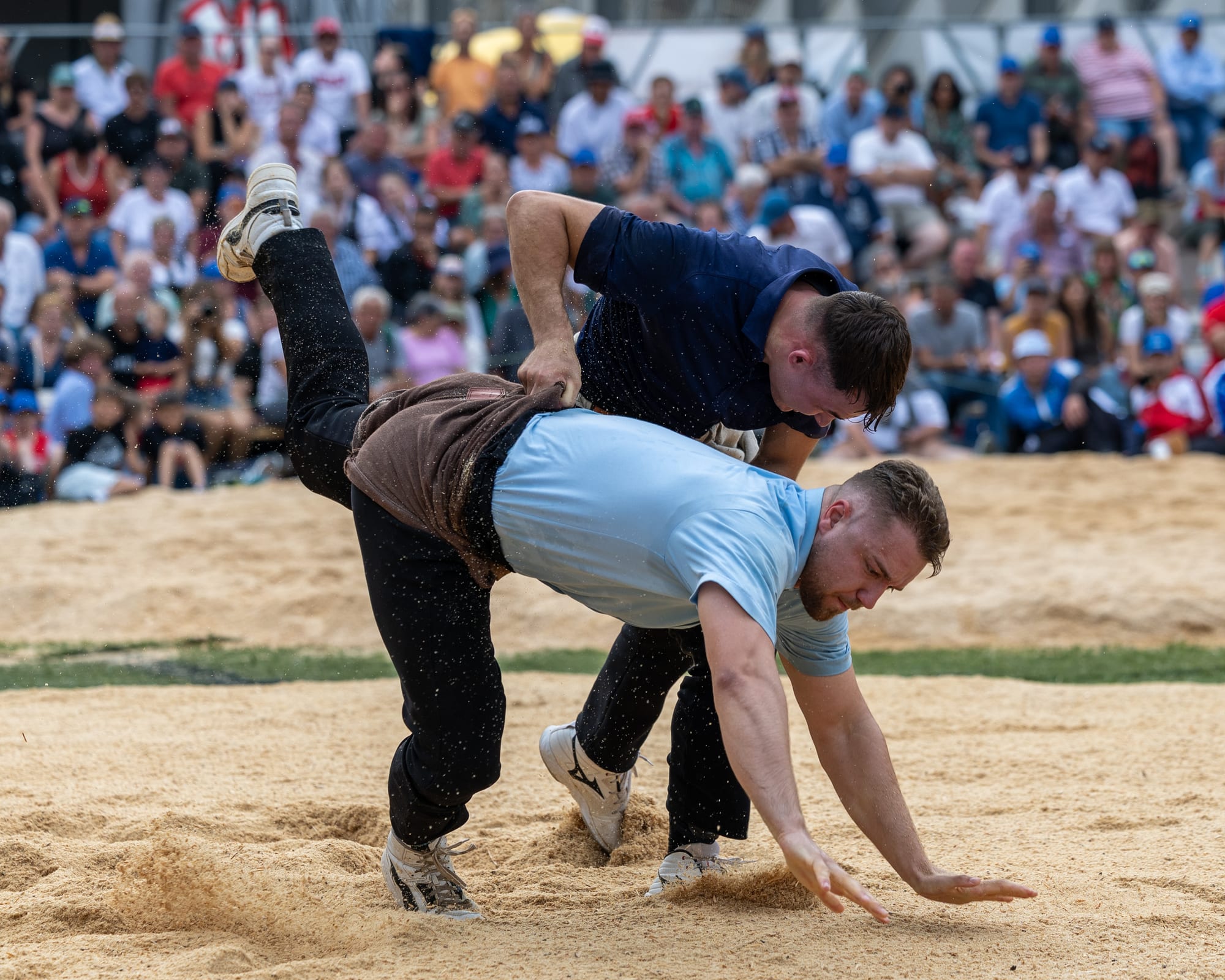 Two wrestlers, one in a dark shirt and one in a light blue shirt, grapple in a ring of sawdust, one lifting the other off his feet by his shorts, in front of a large seated crowd