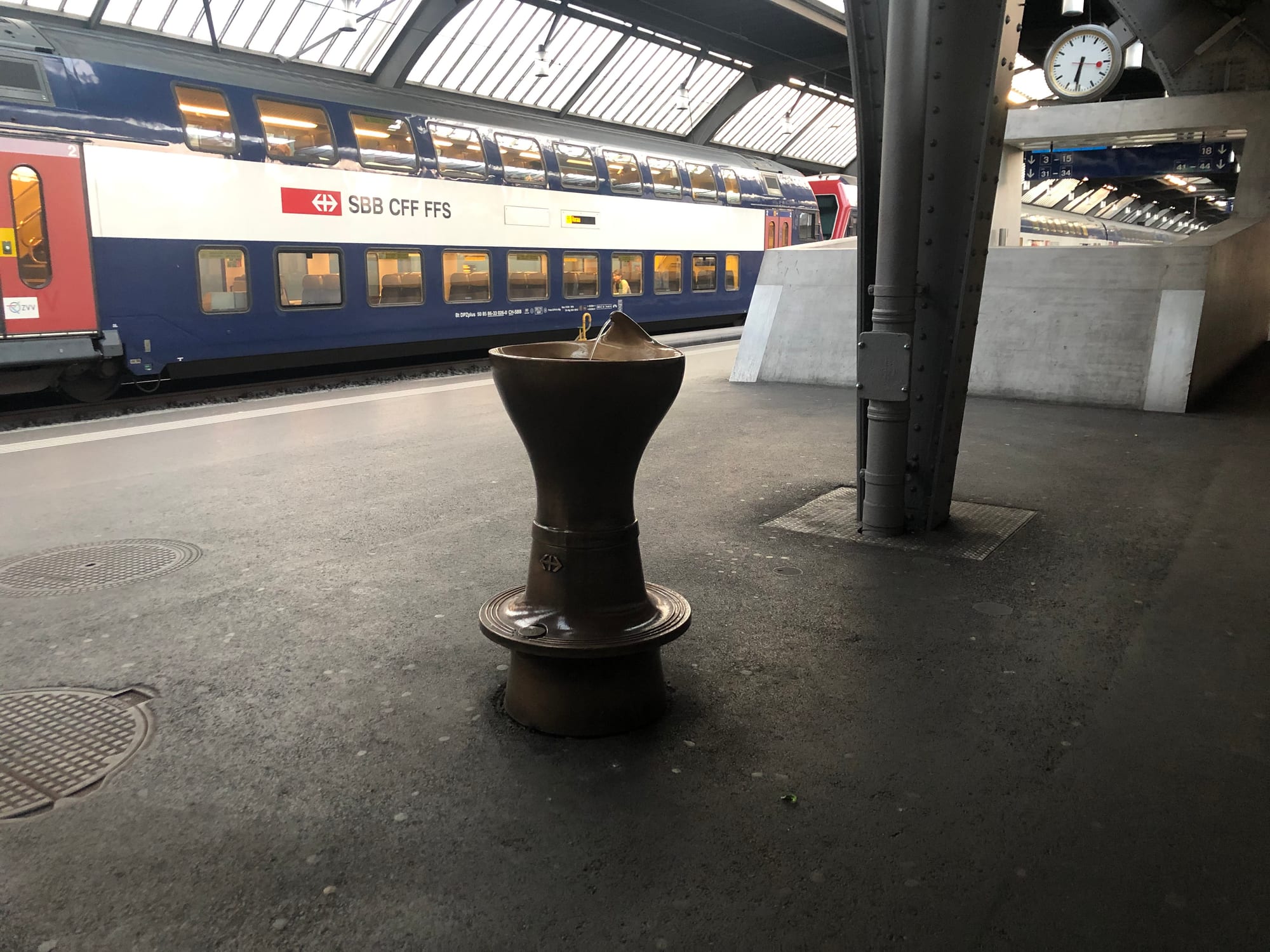 A double-decker Swiss train waiting at a platform in Zurich's main train station, beside a public drinking fountain and a large station clock