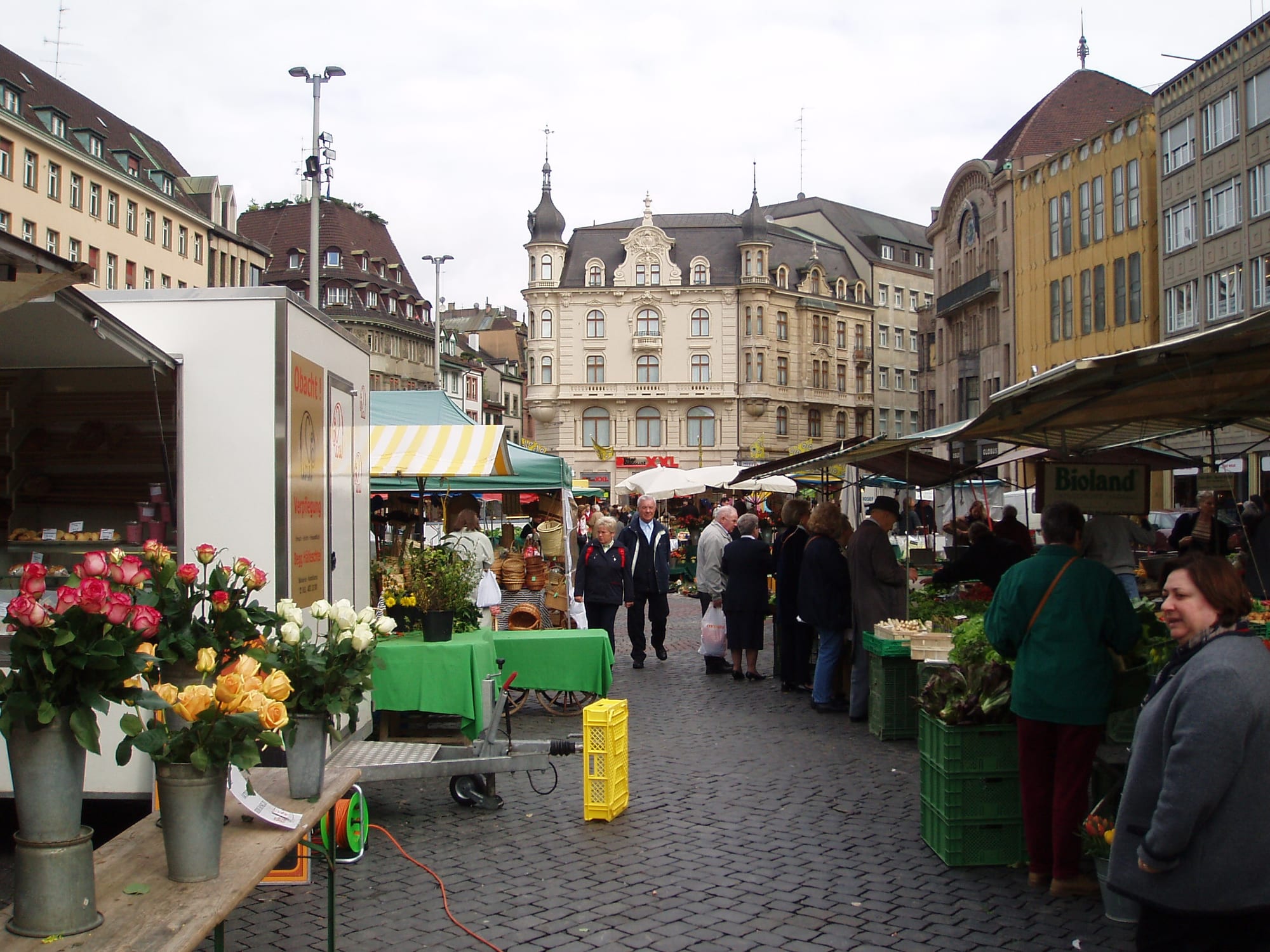 A street market in Basel with flower and produce stalls, market-goers, and historic buildings lining a cobbled square