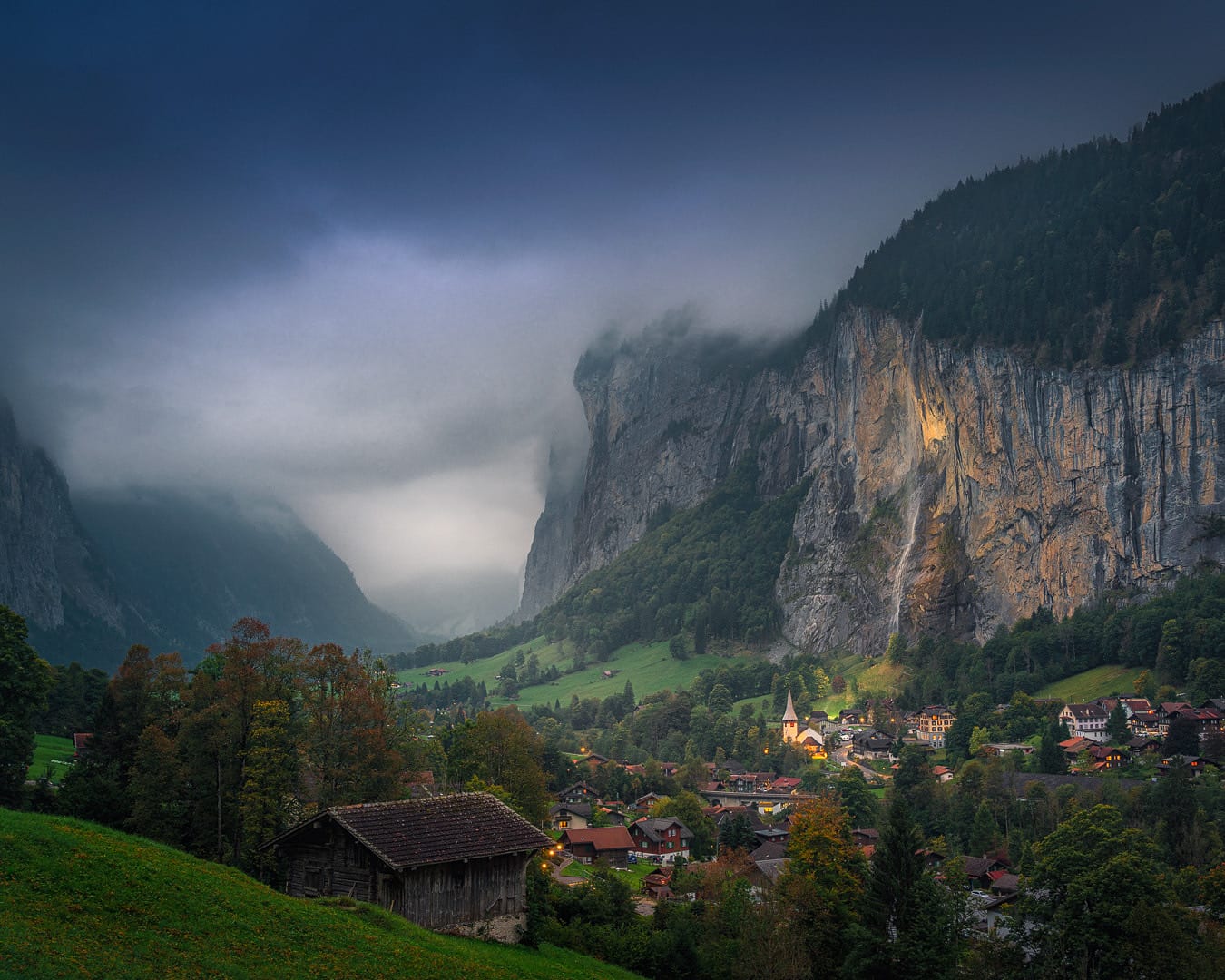 A deep alpine valley with a waterfall streaming off sheer cliffs above a village