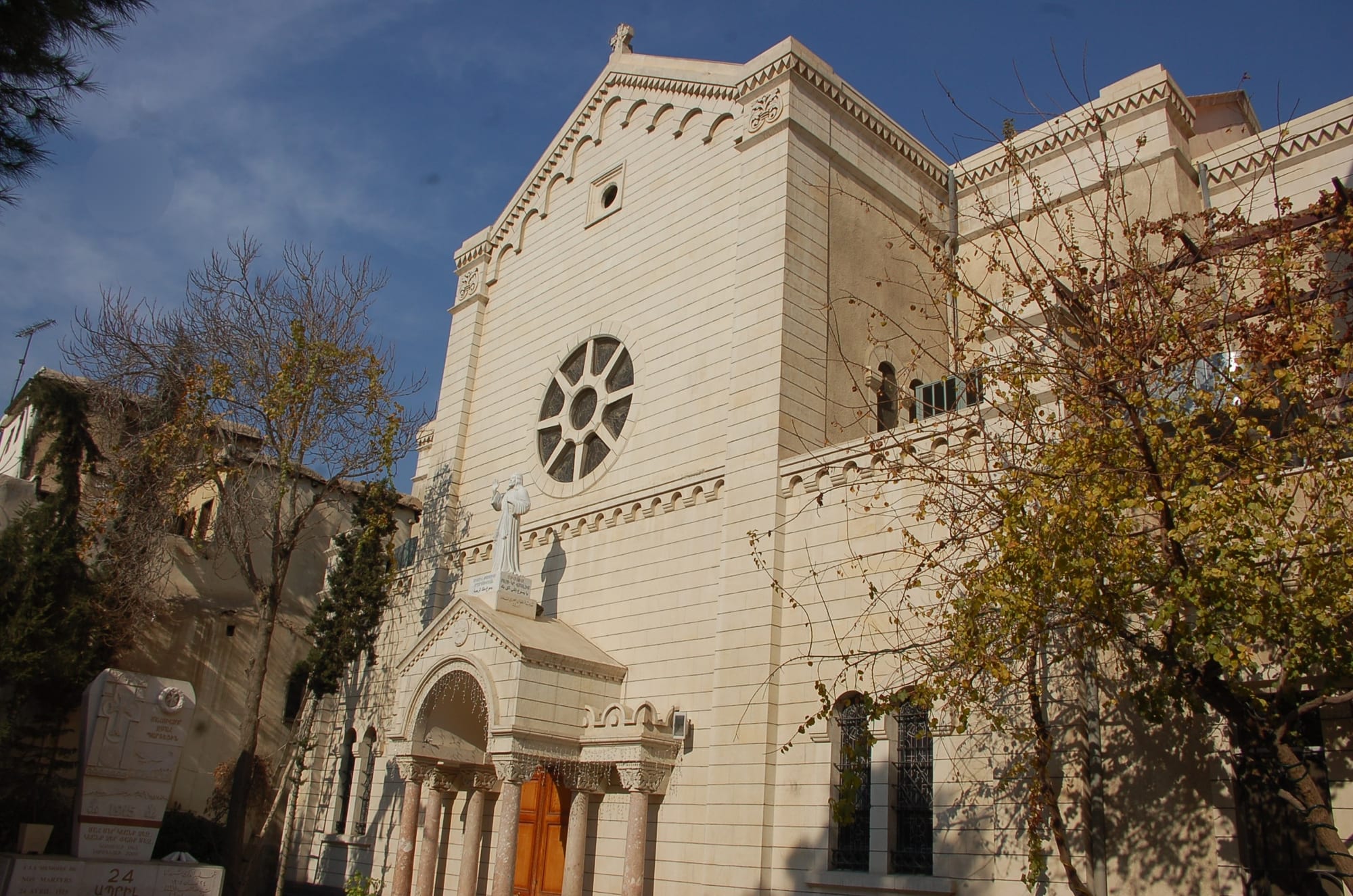 The pale stone facade of a church with a round rose window, a white statue above a columned porch, and autumn trees in front