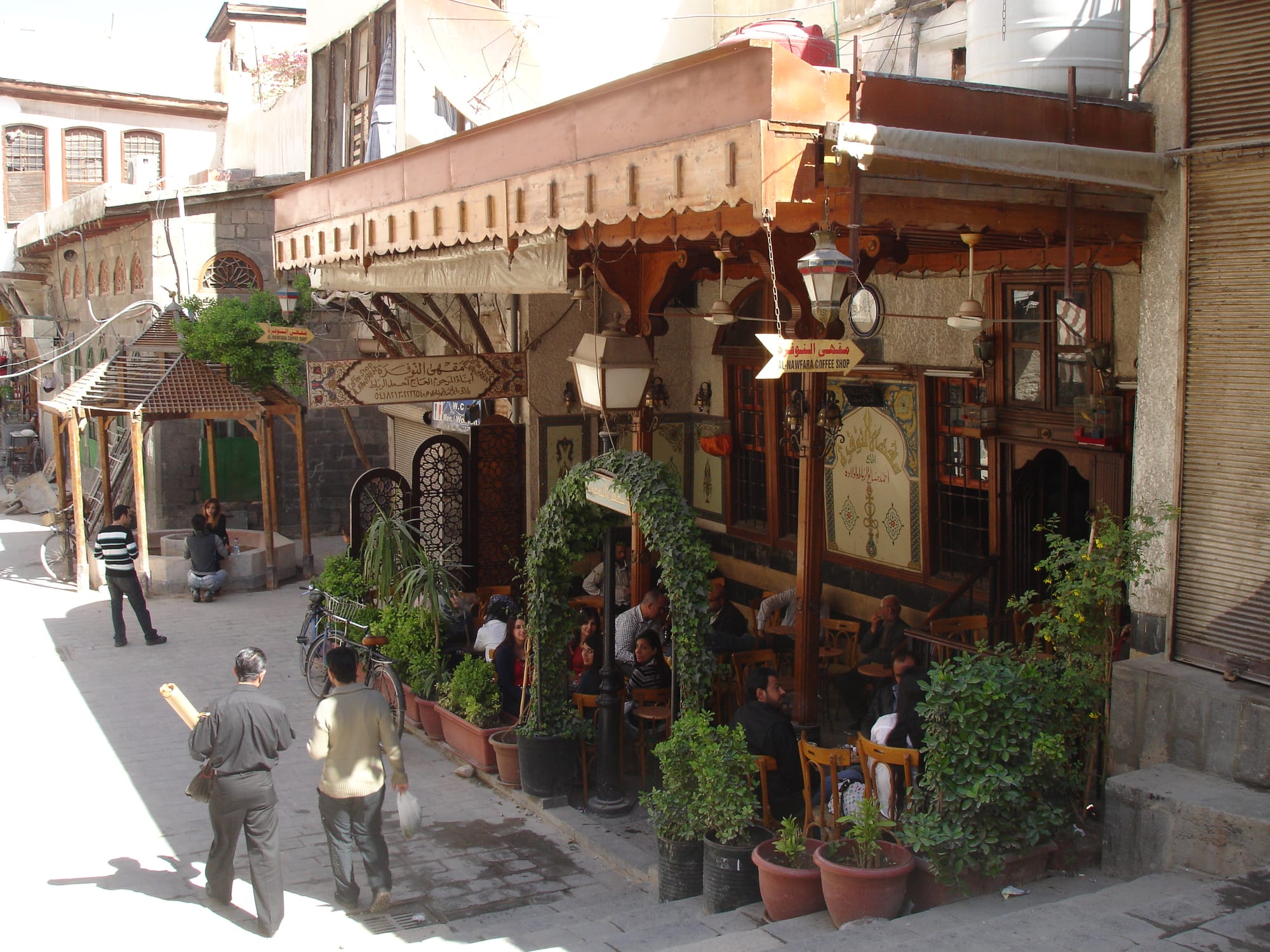 Customers sit at small tables beneath the wooden awning of an old coffee house on a stepped stone street, with passers-by walking past