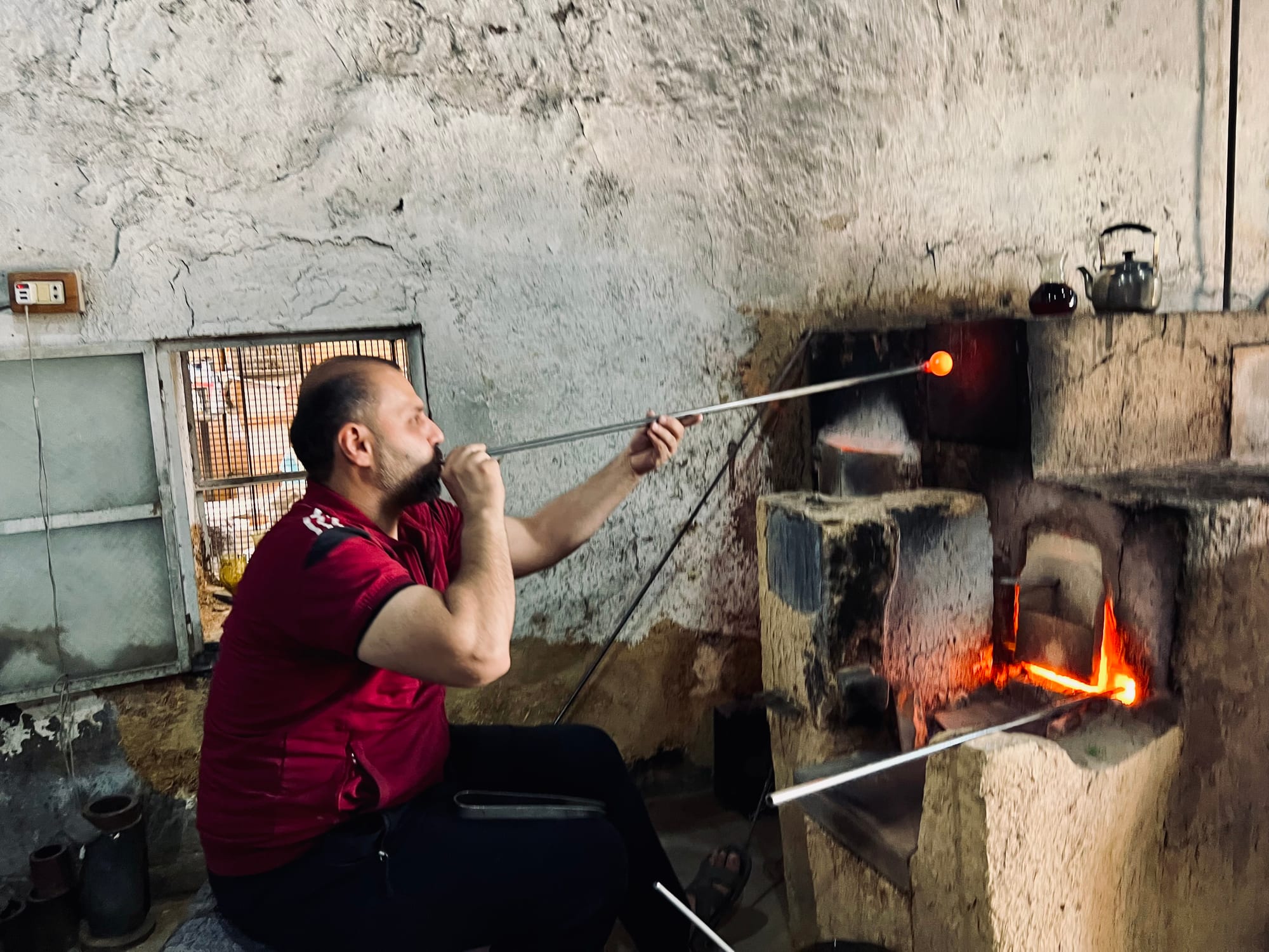 A seated craftsman blows through a long metal pipe with a glowing ball of molten glass at its end, beside a brick kiln with flames visible inside