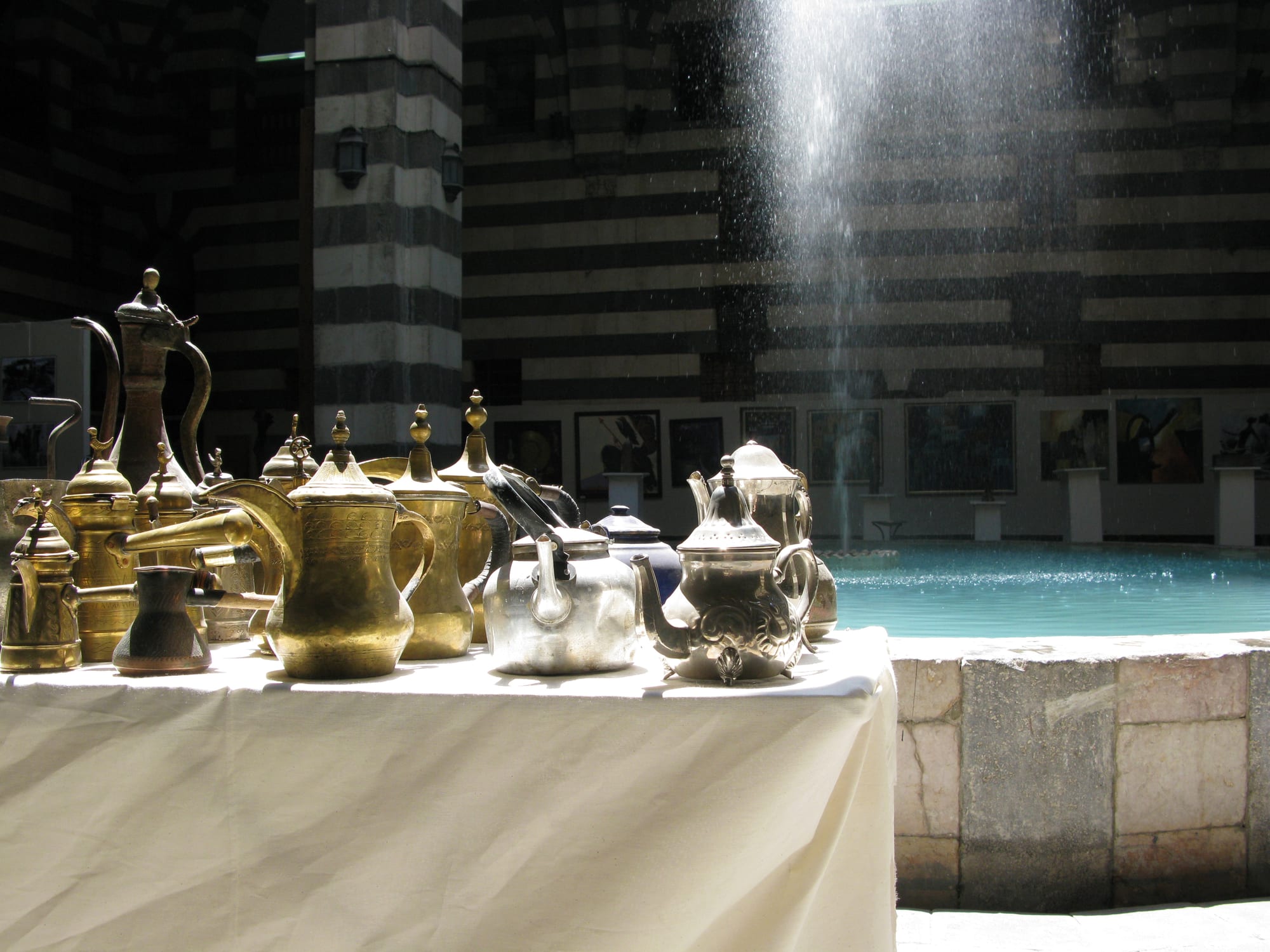 Brass coffee pots with long curved spouts and silver teapots arranged on a white cloth in a sunlit courtyard with striped stone walls and a fountain
