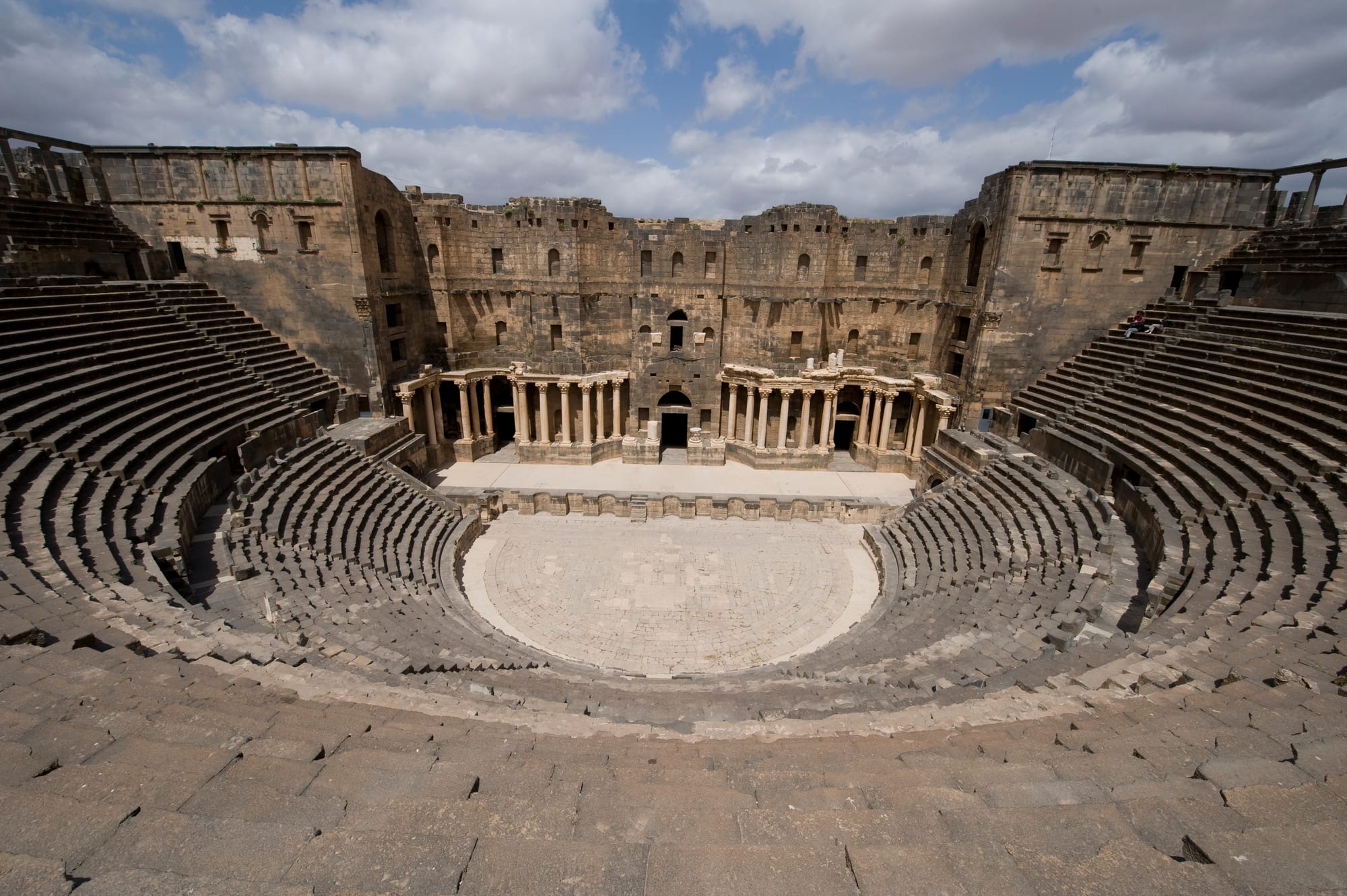 The semicircular tiers of dark stone seating, the paved orchestra and the columned stage wall of the Roman theatre at Bosra