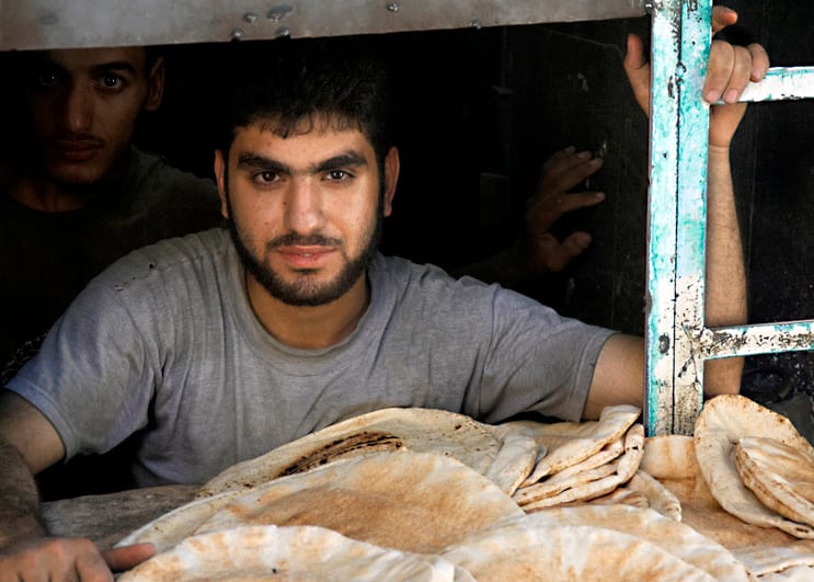 A young man at a bakery hatch holding out fresh flatbread stacked on a metal counter