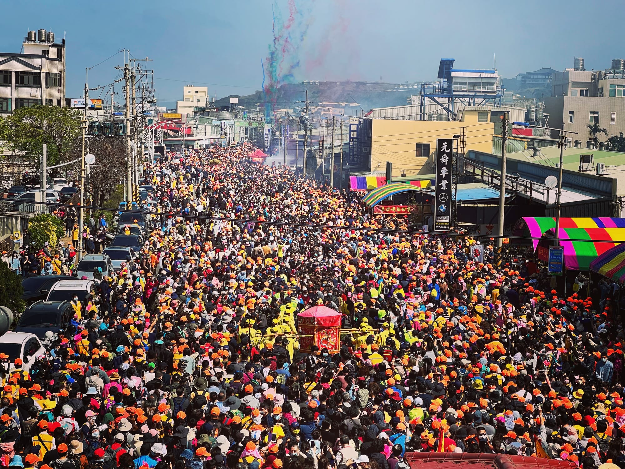 Seen from above, a street packed with thousands of pilgrims, many in orange and yellow caps, surrounds a pink palanquin carried by bearers in yellow, with coloured smoke rising in the distance