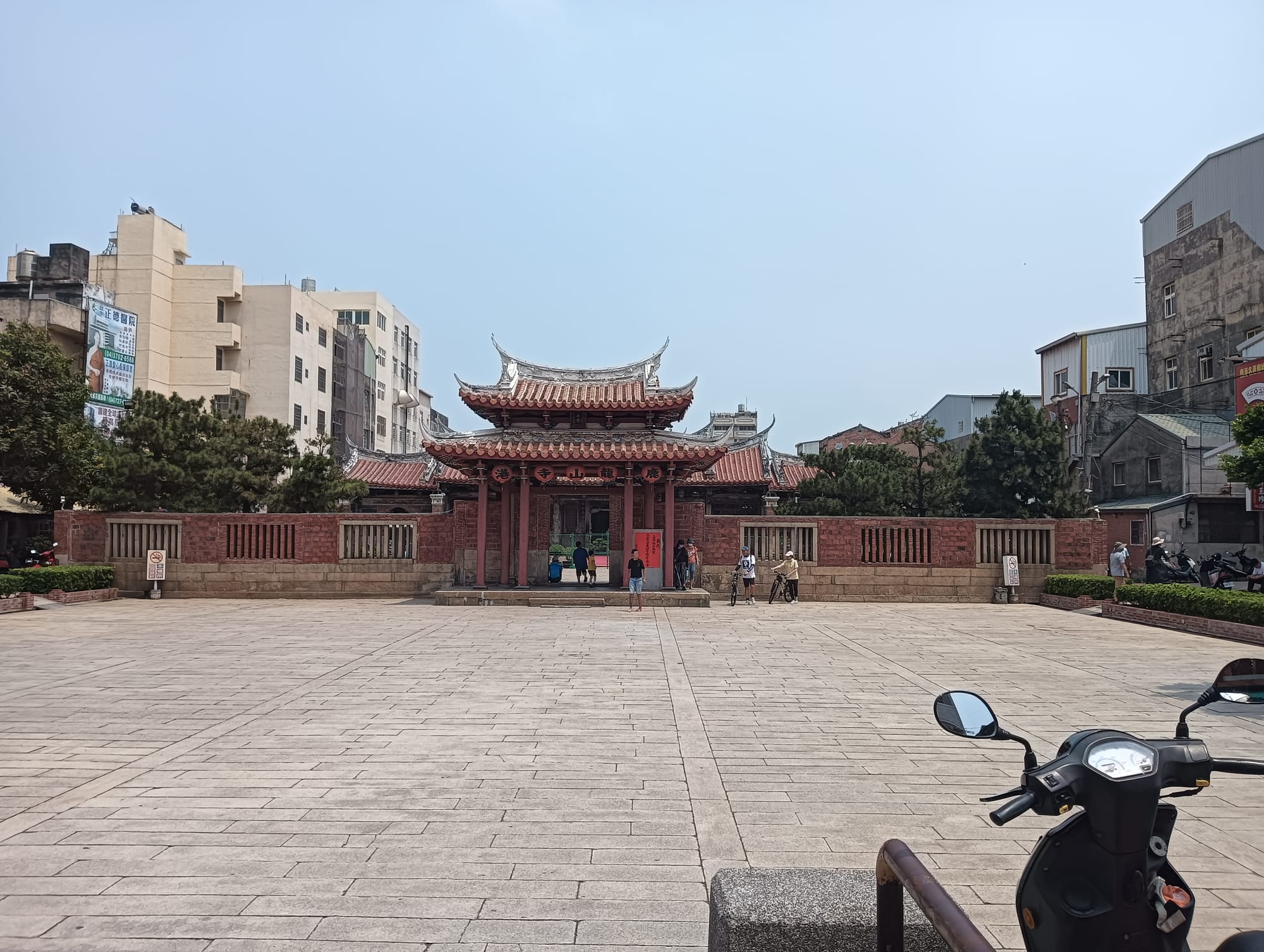 A traditional temple gate with tiered, upswept red-tiled roofs and red columns stands behind a wide paved square, framed by modern apartment buildings