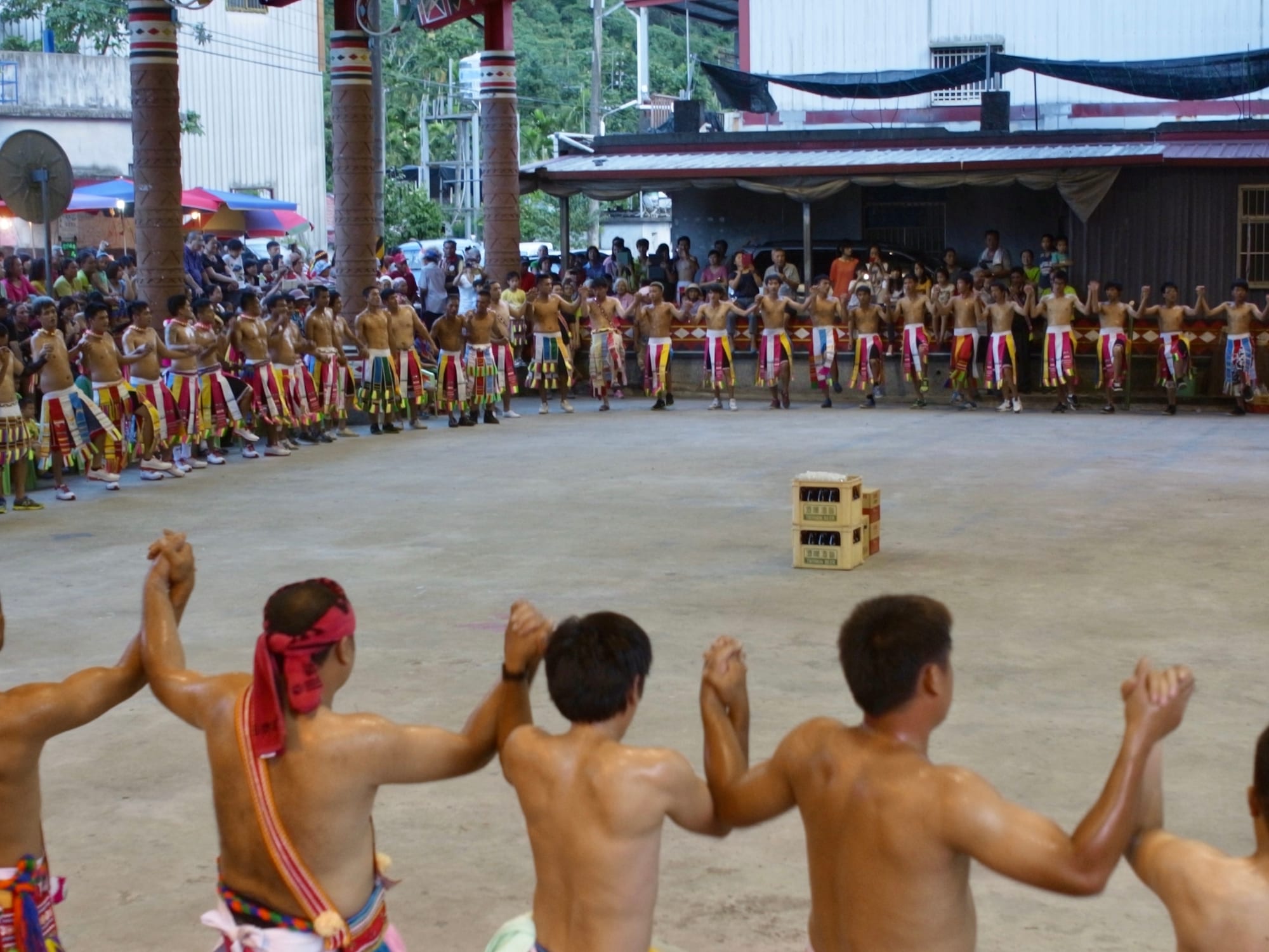 Rows of young men in colourful woven skirts and headbands, hands linked and raised, dance in a large circle on an open concrete ground, with spectators behind