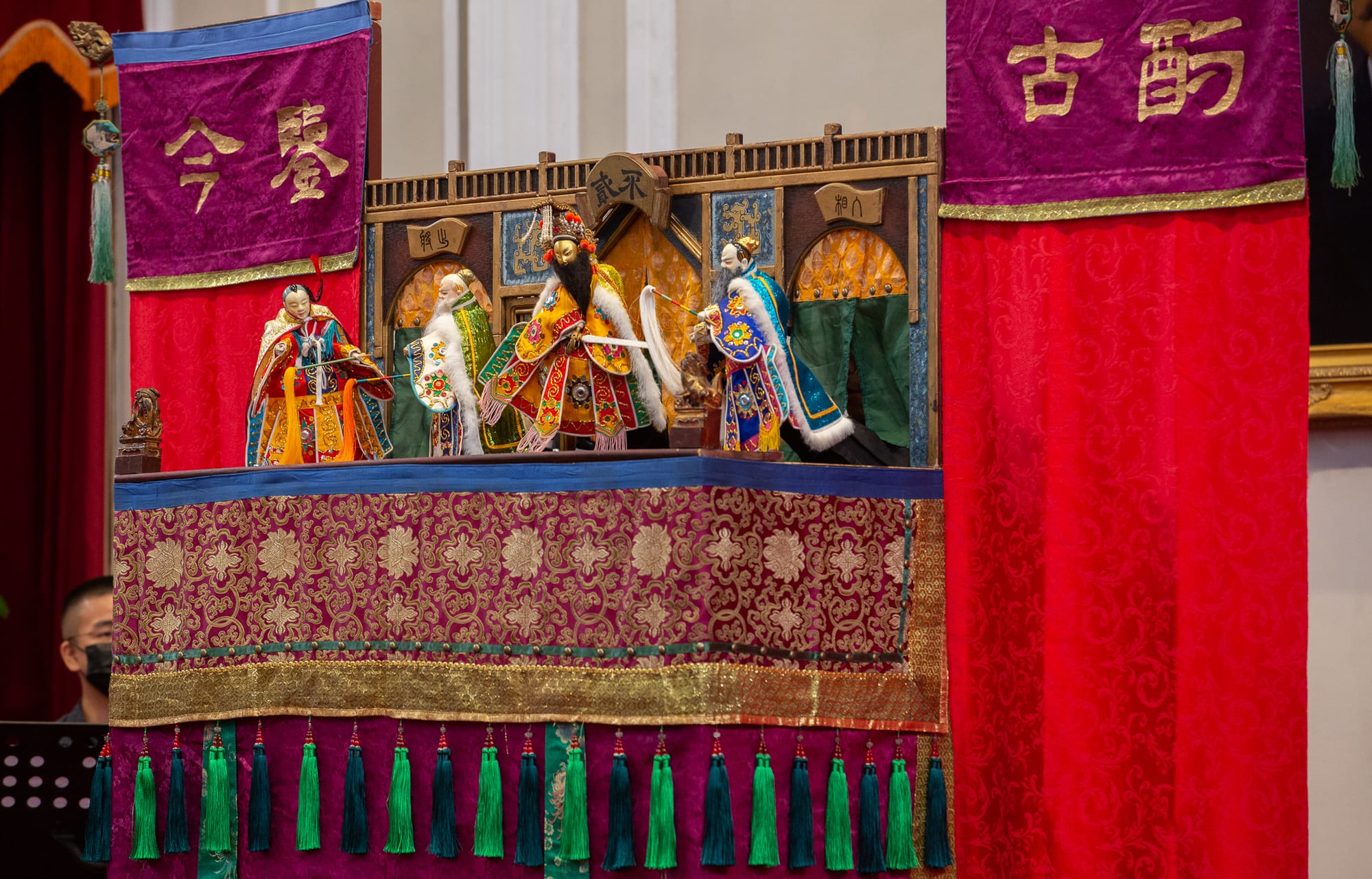Four glove puppets in elaborate embroidered robes and headdresses stand on a small carved and gilded puppet stage draped with purple and red embroidered cloth