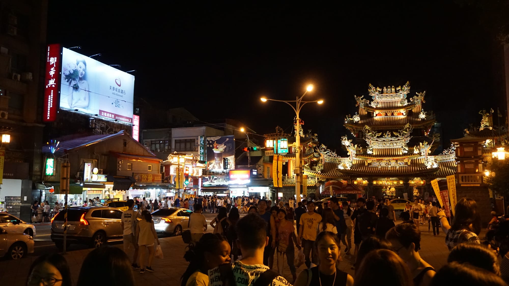 An illuminated temple with an ornate tiered roof beside a crowded night market street lined with stalls and neon signs