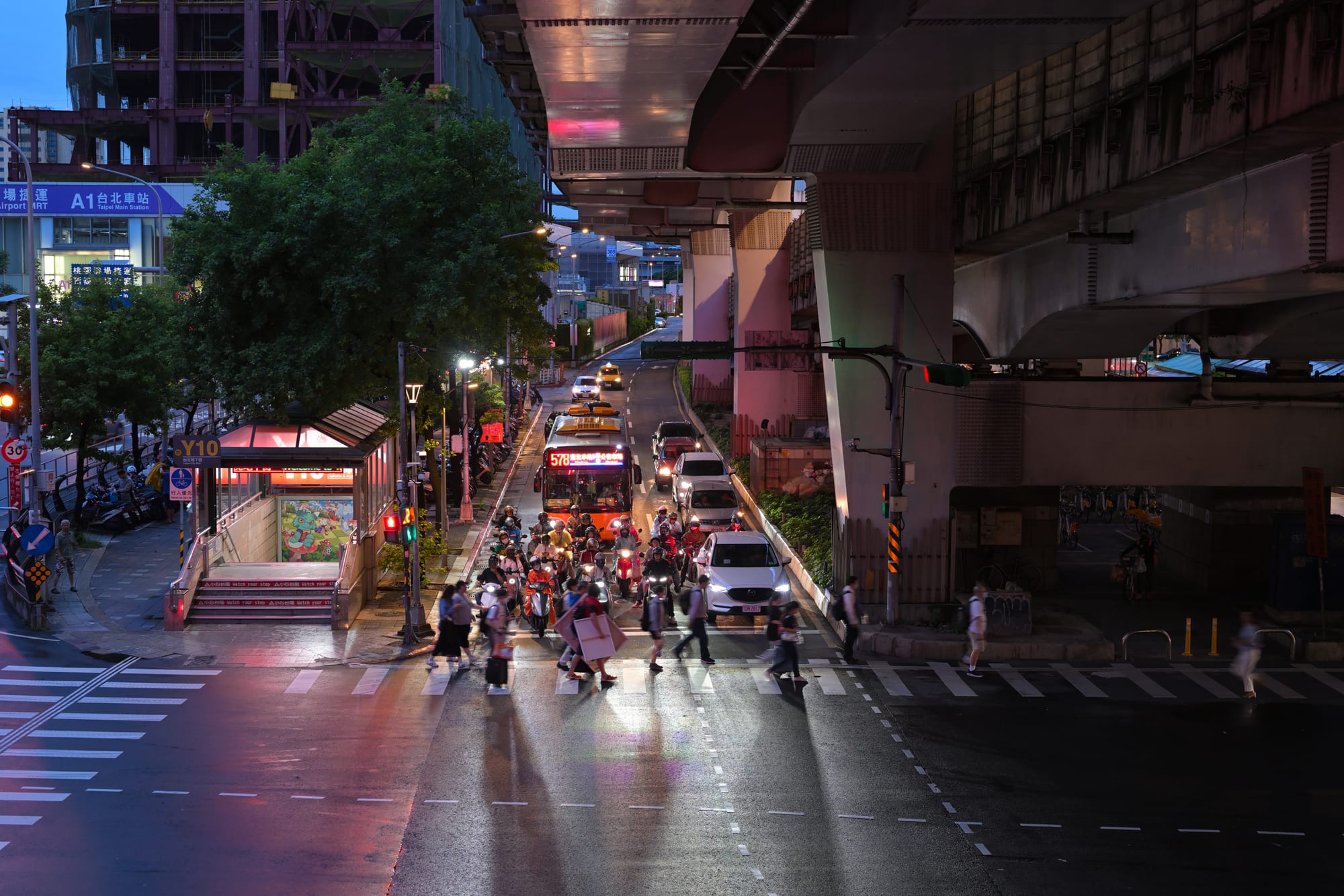 A dusk street scene near Taipei Main Station with scooters and pedestrians crossing under an elevated road and glowing shopfronts