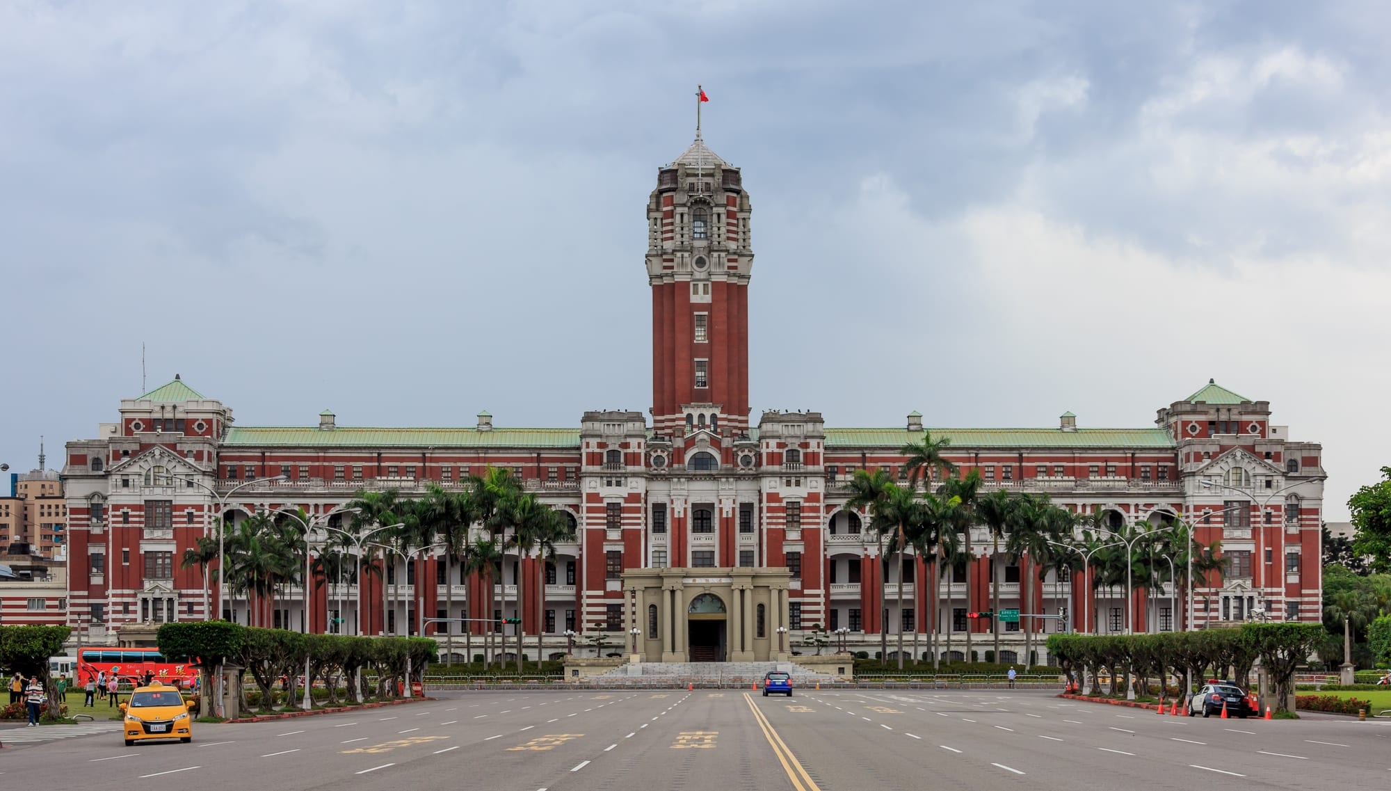 A long red-and-white building with a tall central tower flying a flag, seen head-on across a wide empty boulevard lined with palms