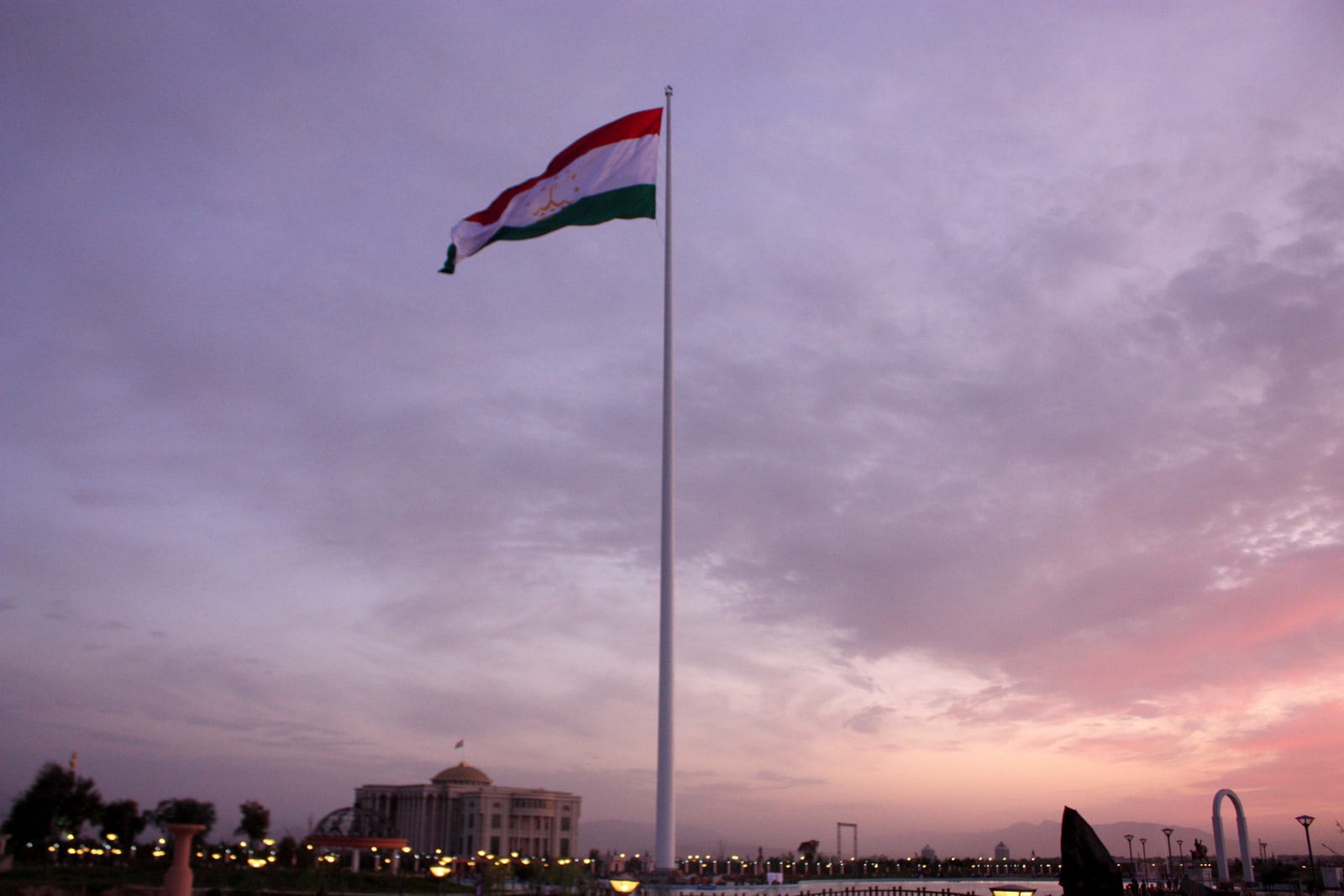 A very tall white flagpole flying the red, white and green Tajik flag over a landscaped park at dusk, with the presidential palace lit up in the background