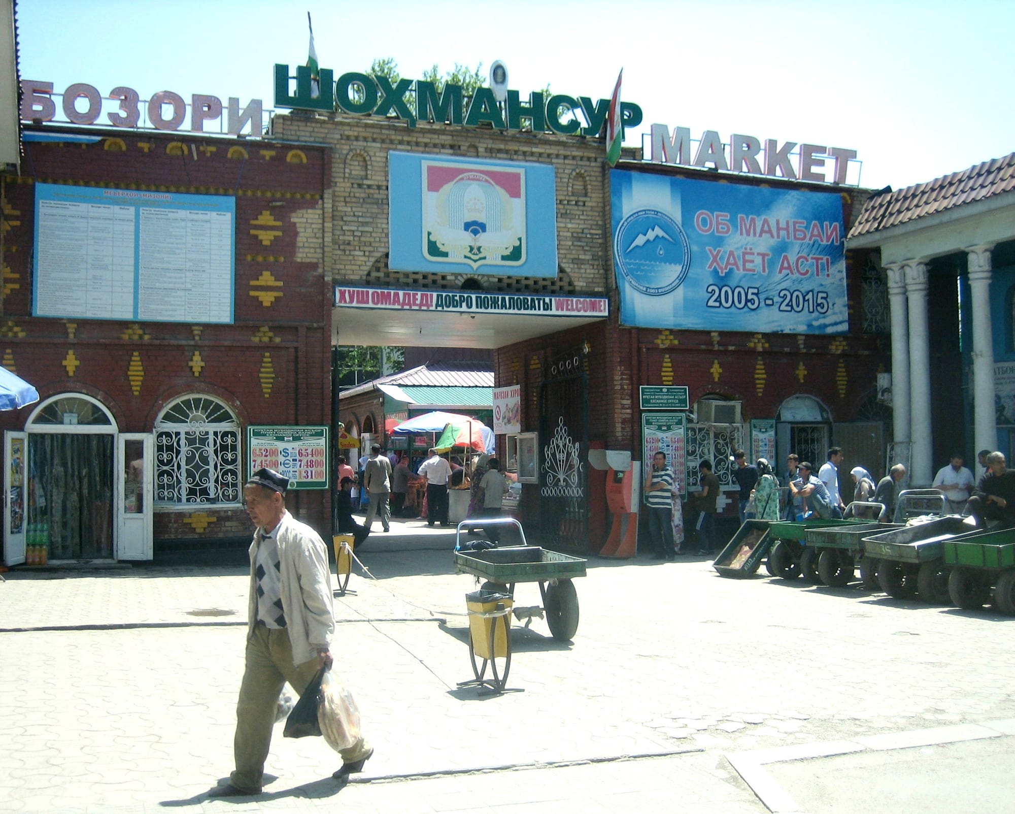 The brick entrance gate of Shohmansur Market in Dushanbe, decorated with signage in Tajik, Russian and English, with shoppers and handcarts passing through