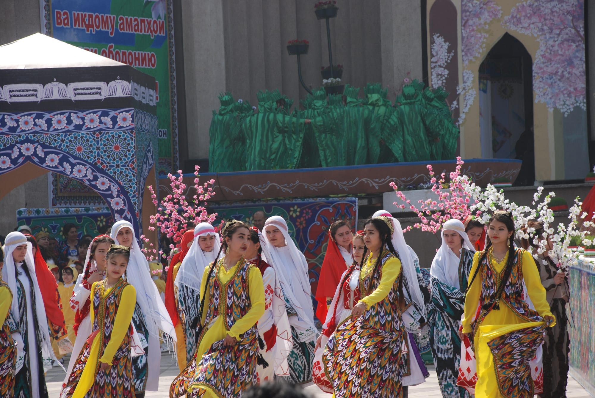 Rows of young women in yellow and brightly patterned dresses, some with white or red headscarves, walking in procession in front of a stage hung with blossom branches