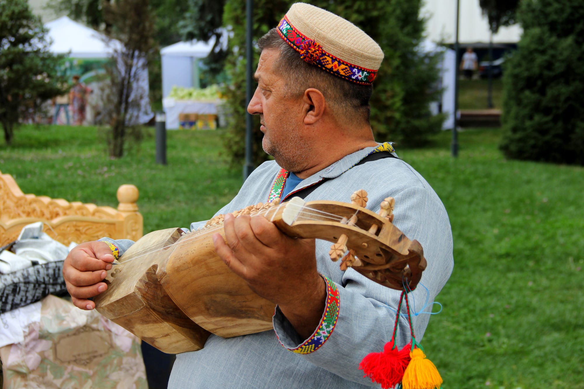 A man in a pale blue shirt with an embroidered collar and a woven skullcap with a bright embroidered band holds a carved wooden stringed instrument with red and yellow tassels hanging from its neck