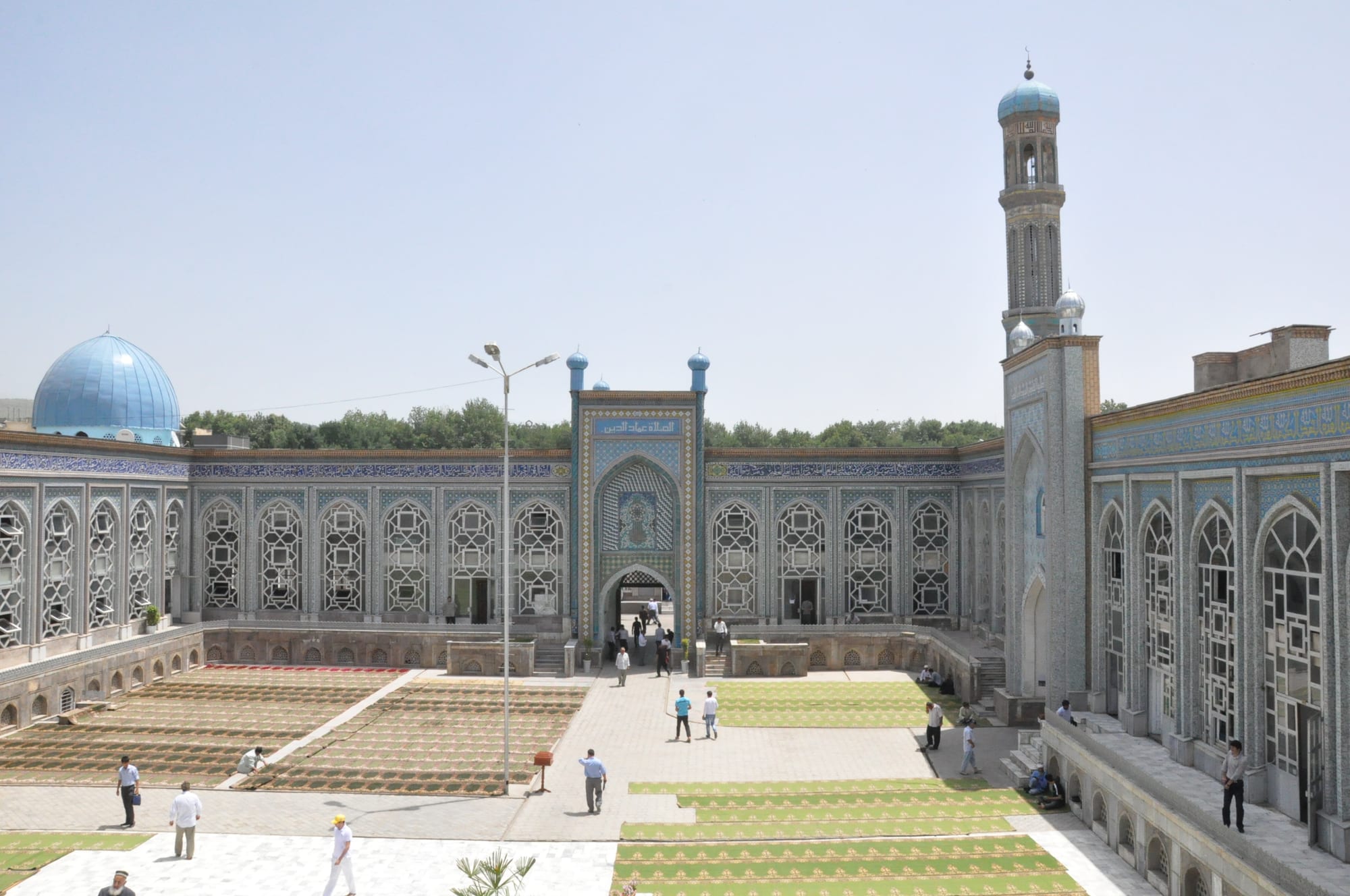 A wide tiled courtyard enclosed by arcades decorated with blue and white tilework, with a turquoise dome on the left, a tall minaret on the right and rows of prayer rugs laid out on the paving