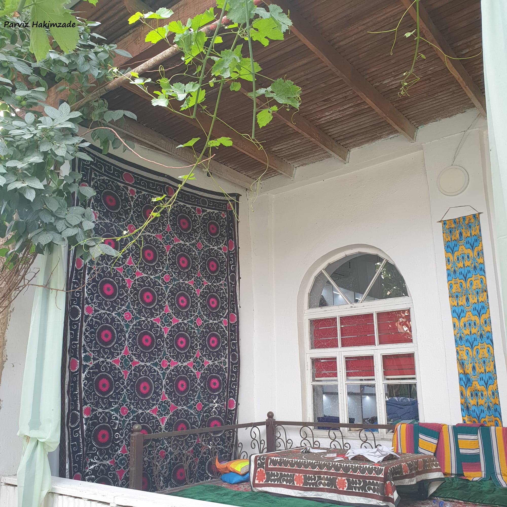 A shaded veranda of a white house with a large black and pink embroidered hanging on one wall, a patterned silk panel beside an arched window, and a low table surrounded by cushions and striped bolsters under a vine