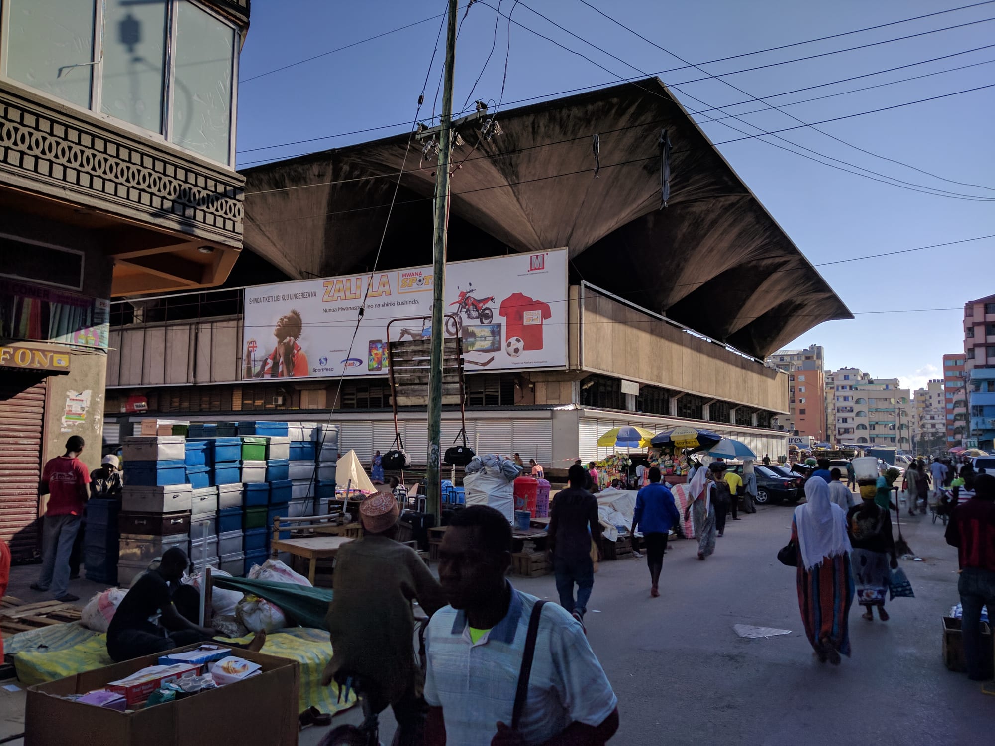 A busy street market at dawn in front of a large covered market hall, with vendors setting up stalls and pedestrians walking past