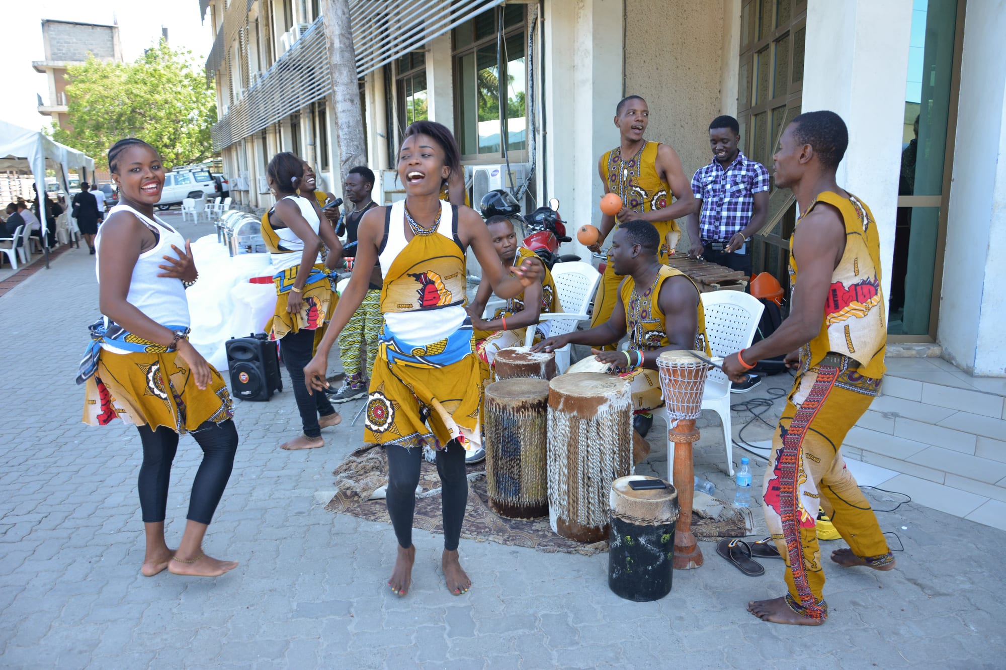 Young women in yellow and white patterned costumes dancing and singing on a paved forecourt beside a group of tall wooden drums, with male musicians in matching yellow behind them
