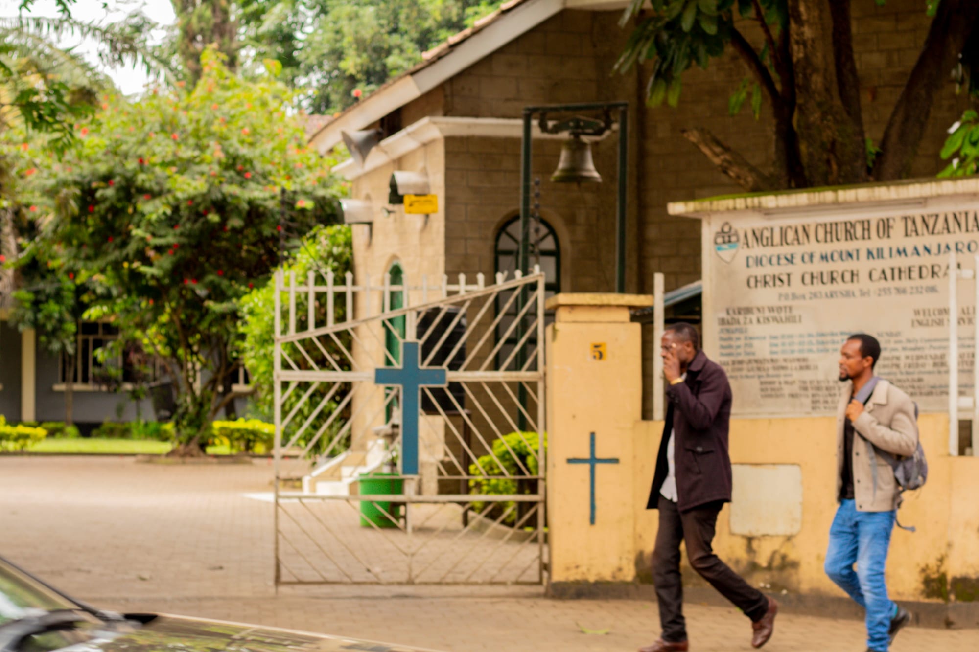 Two men in jackets walk along a pavement past the gate of a stone church, with a blue cross on the gate, a bell hanging above the entrance and a sign reading Anglican Church of Tanzania