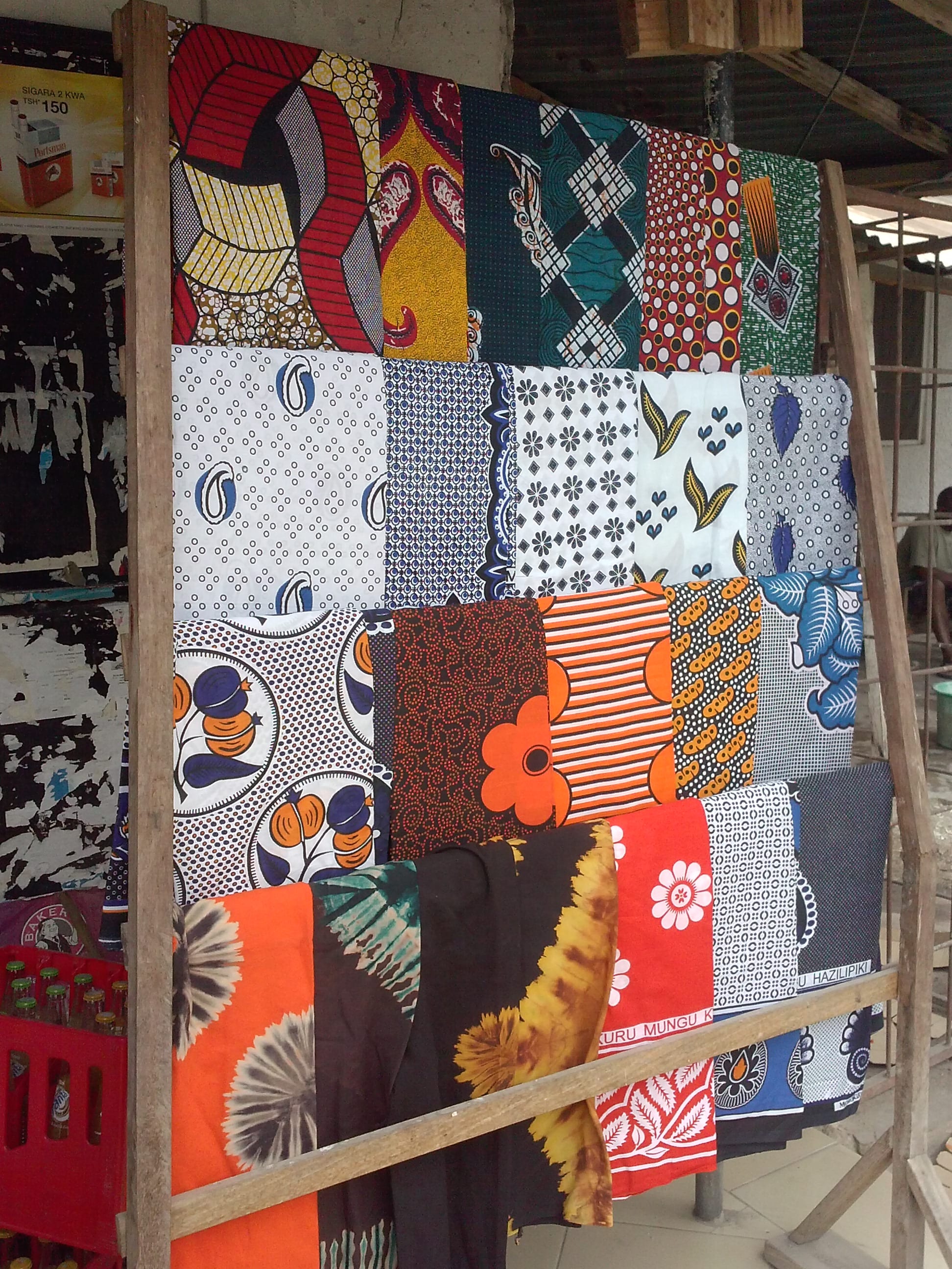 A wooden rack hung with rows of brightly printed cotton cloths in red, orange, blue, black and white patterns, some with lettering printed along the hem