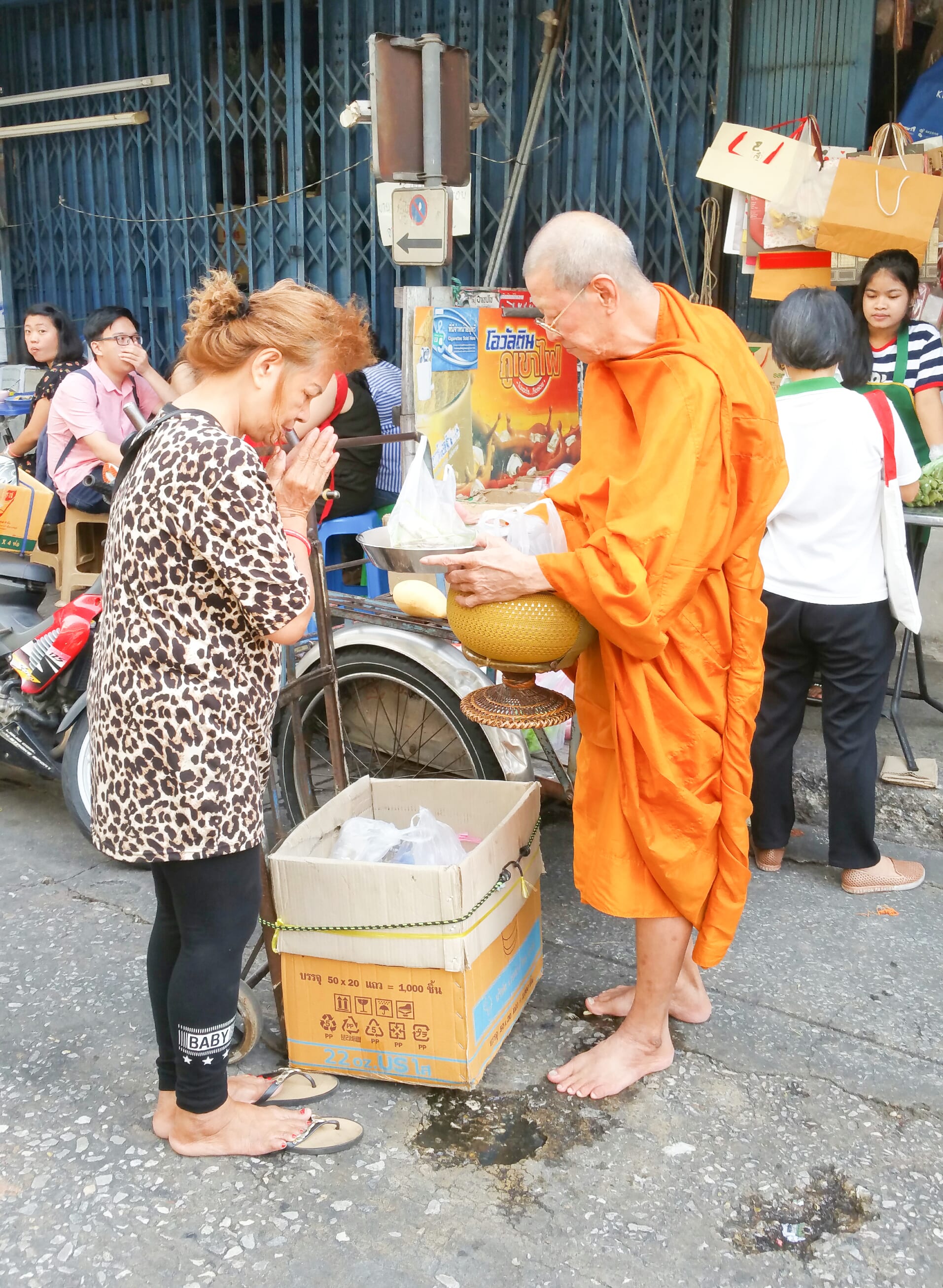 A woman with hands pressed together offering food to a barefoot Buddhist monk in orange robes on a city street