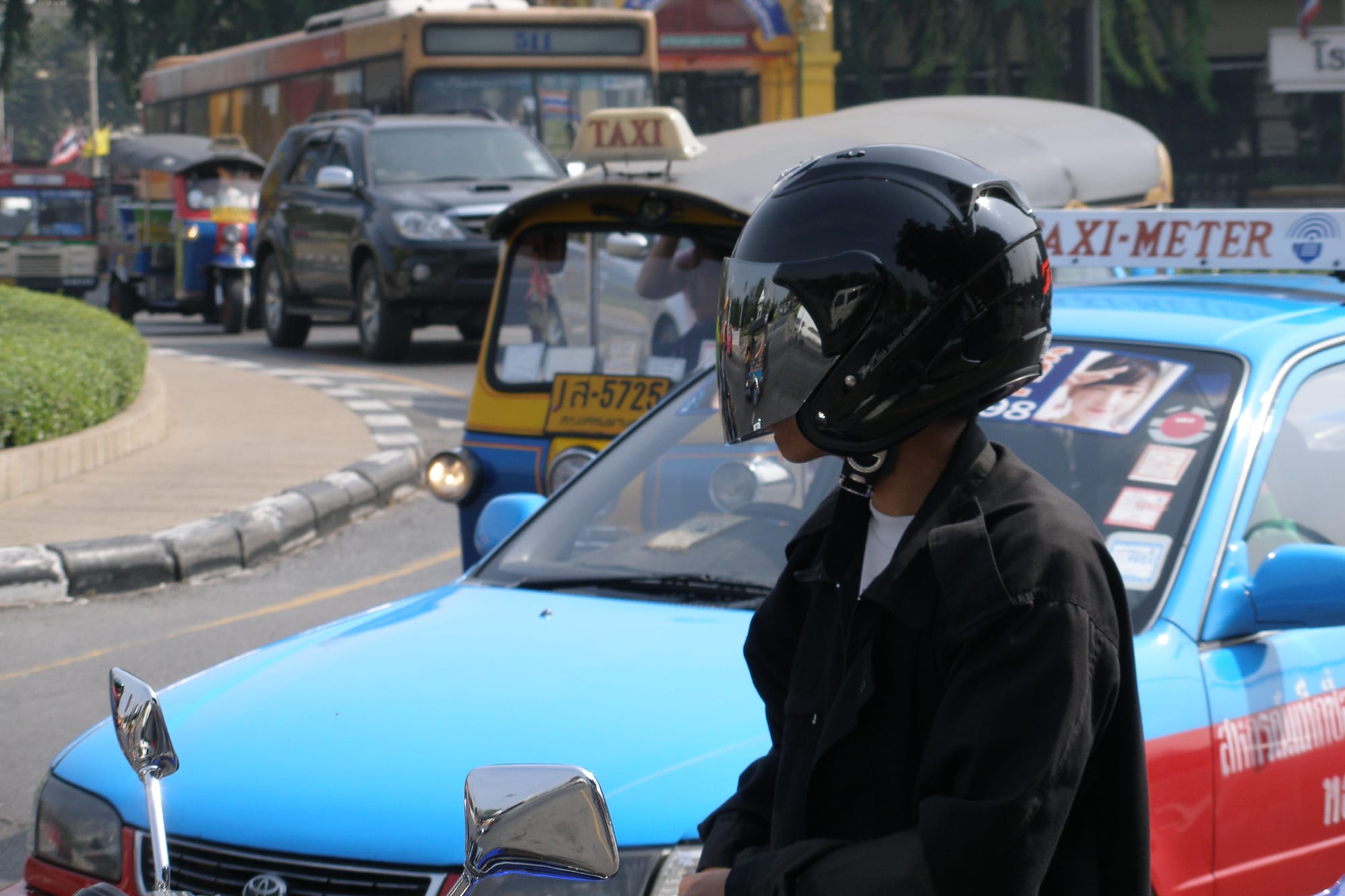 A Bangkok street scene with a motorbike rider in the foreground and taxis, a tuk-tuk and a bus in traffic behind