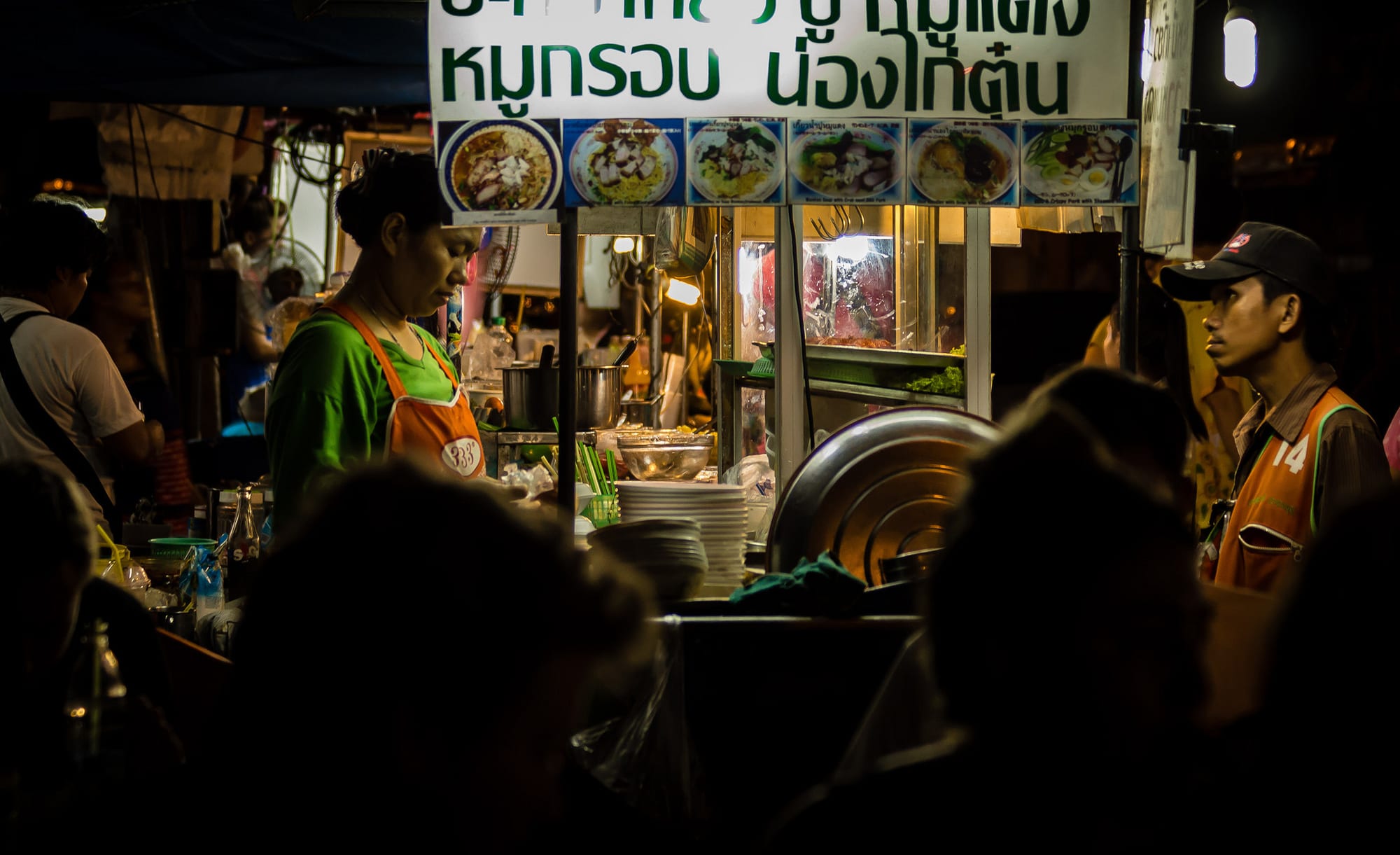 A street food vendor serving customers at an illuminated stall with a menu board of noodle dishes