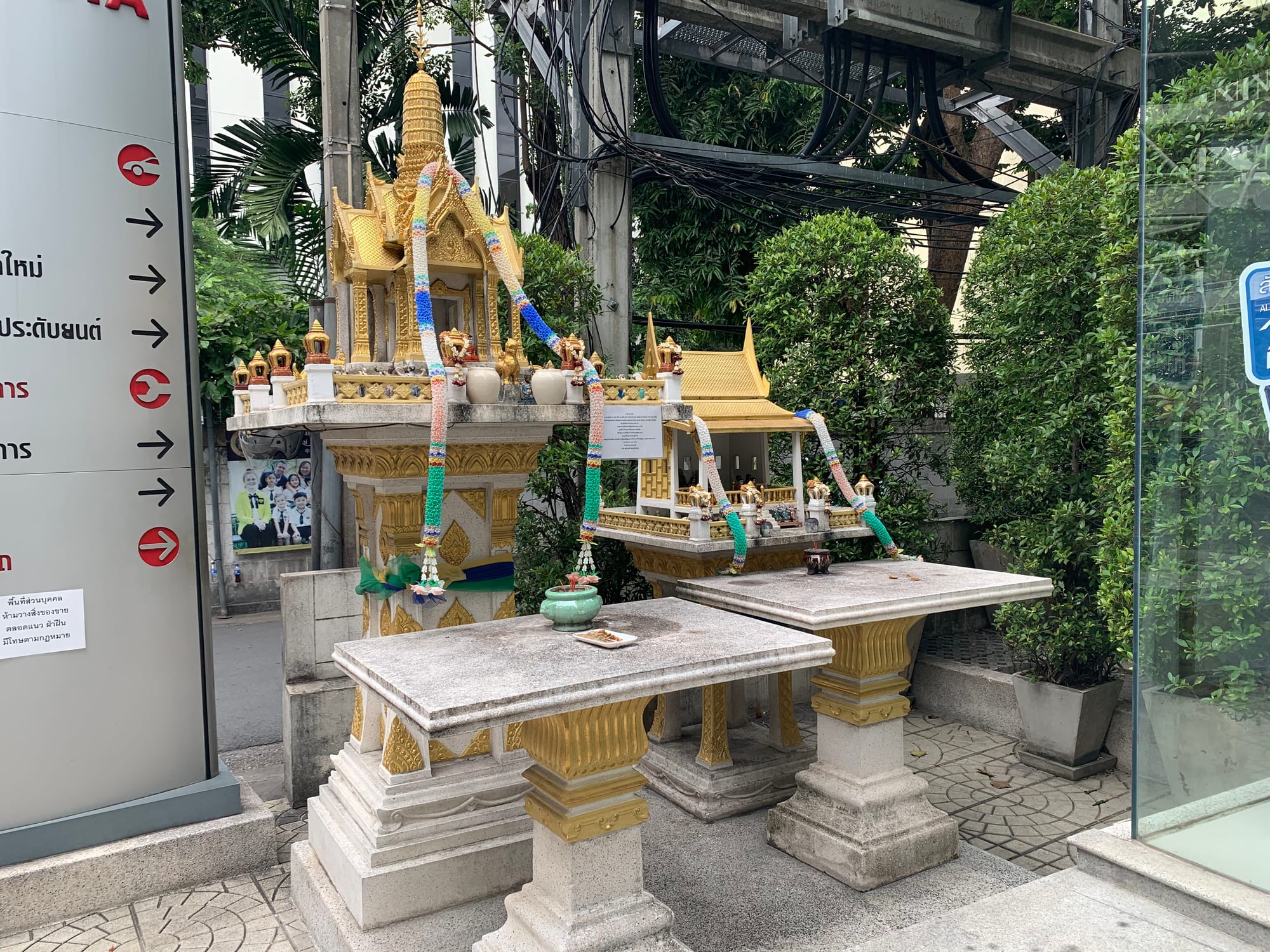 Two ornate gilded spirit houses on white pillars beside a city pavement, hung with long garlands of coloured flowers and set with small figures, vases and offerings, in front of trees and overhead cables