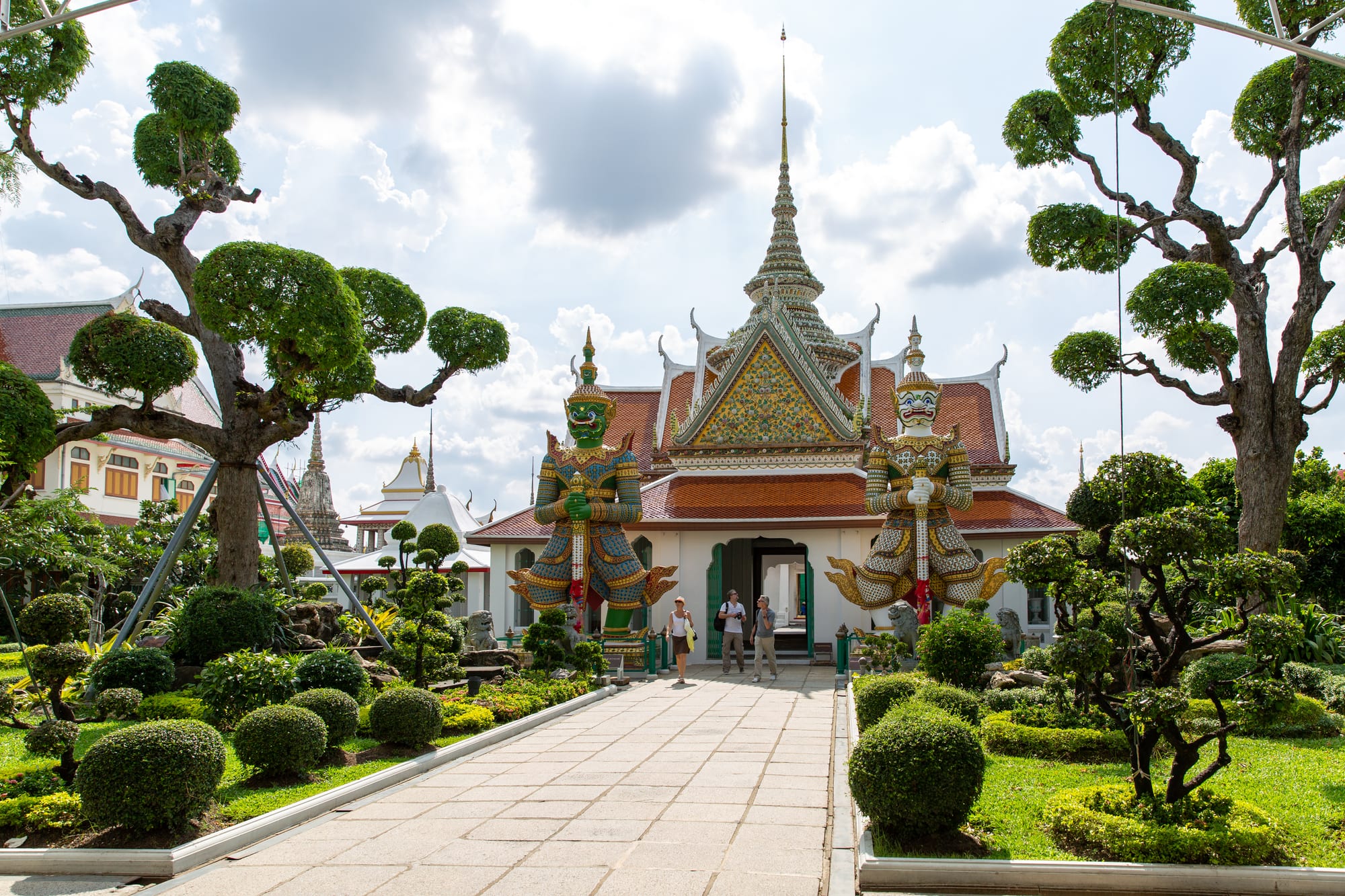 A paved path between clipped trees and shrubs leading to a white temple hall with tiered red and green roofs, a tall spire and two giant painted guardian statues flanking the doorway