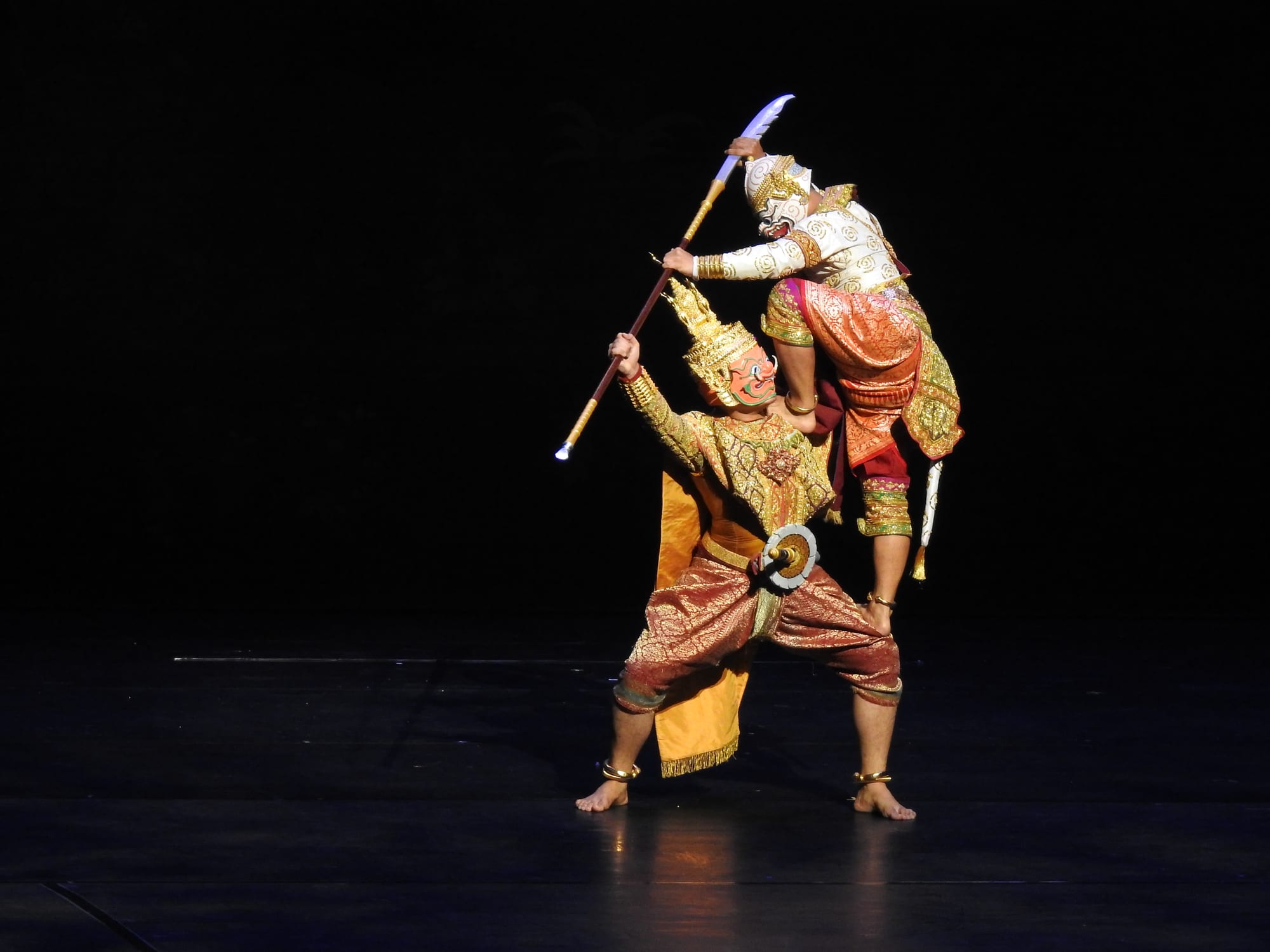 Two khon performers in masks and gold embroidered costumes on a dark stage, one standing on the thigh of the other and raising a halberd above his head