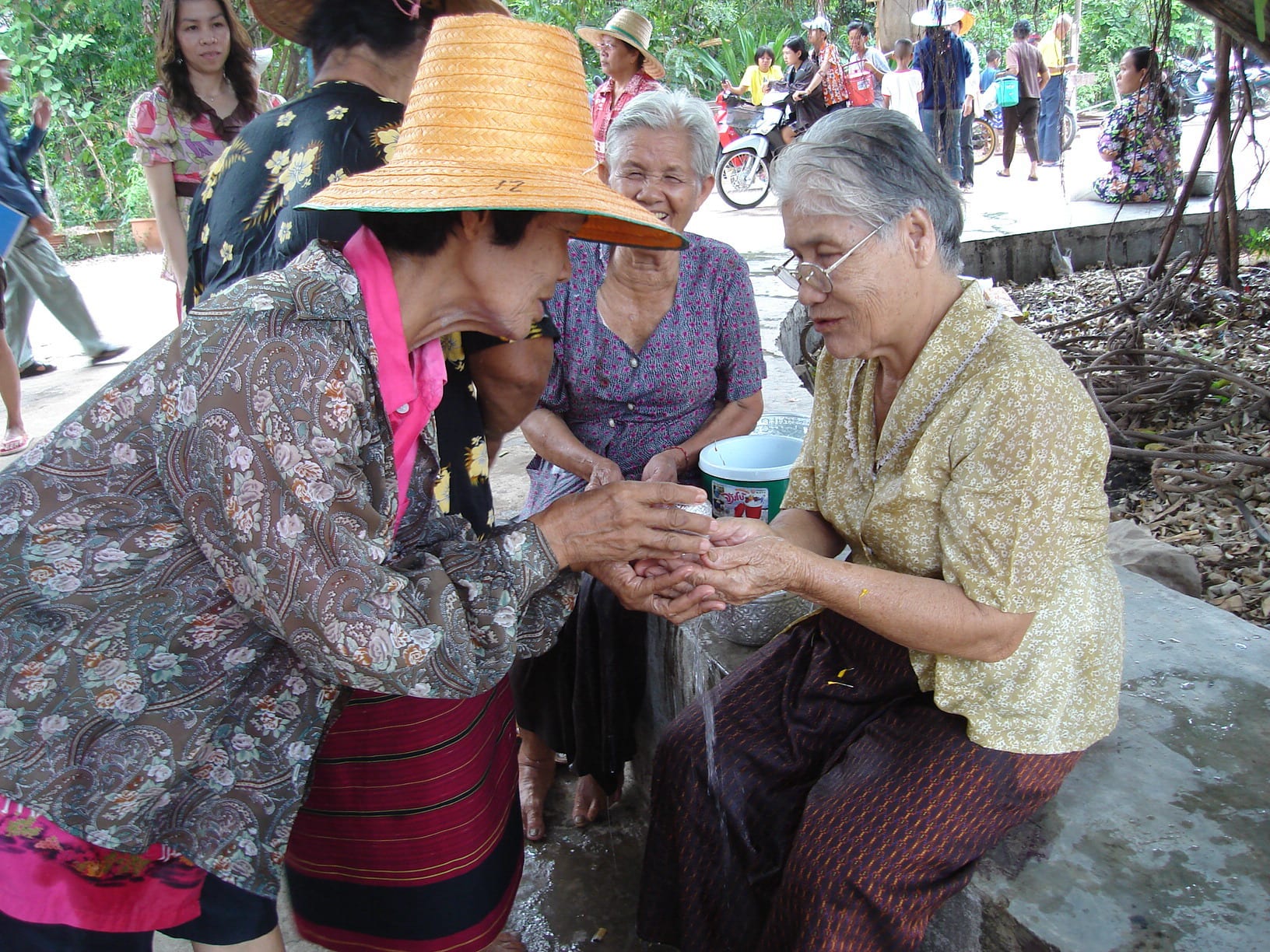 An older woman in a straw hat pours water from a small bowl over the cupped hands of a grey-haired woman seated on a low wall, while other women in patterned blouses look on