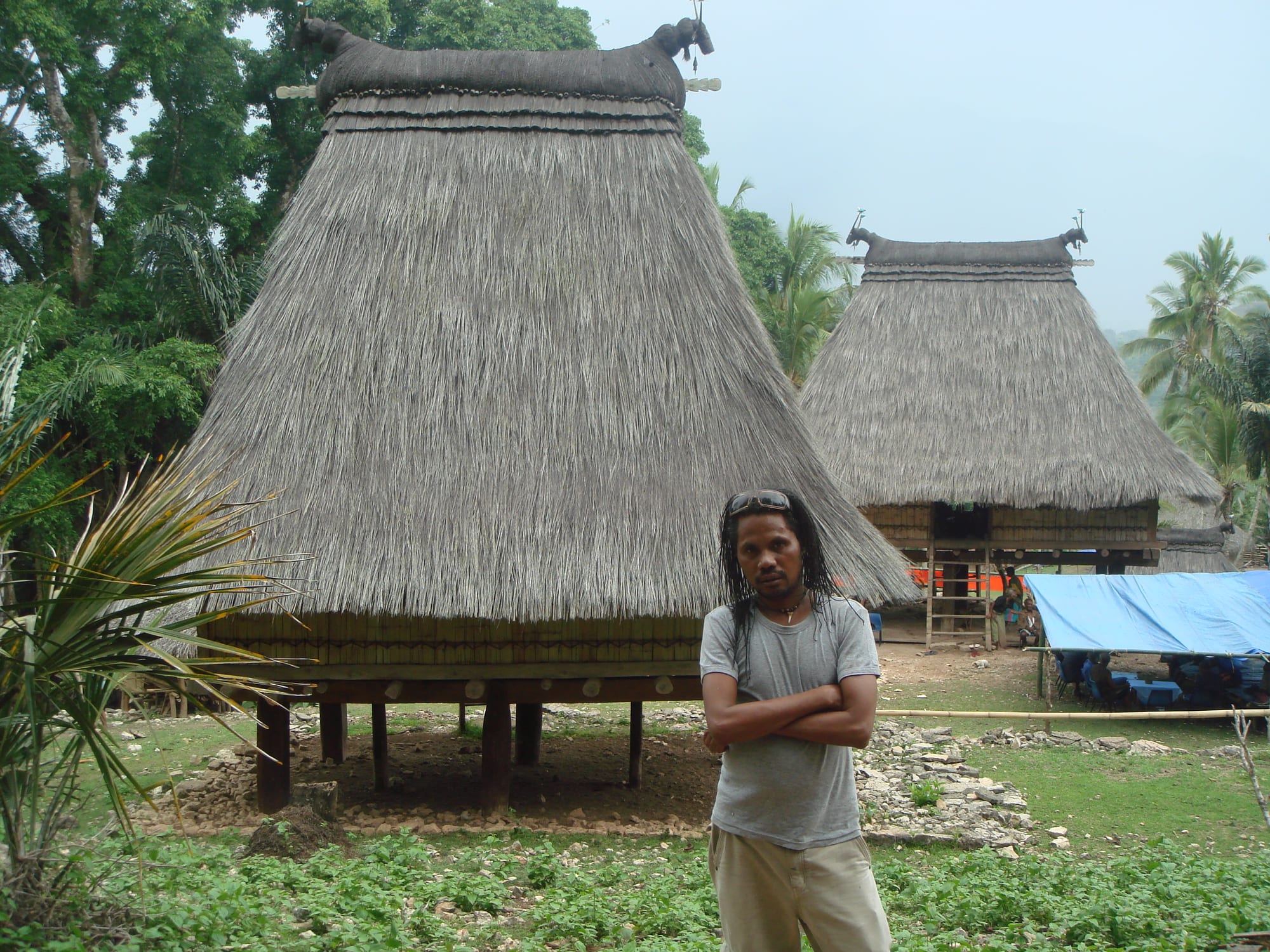 Two tall, steeply pointed thatched-roof houses raised on stilts, with a person standing in front of them