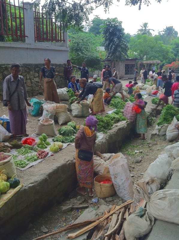 Vendors and customers at an outdoor vegetable market, with produce laid out on the ground and low stone walls