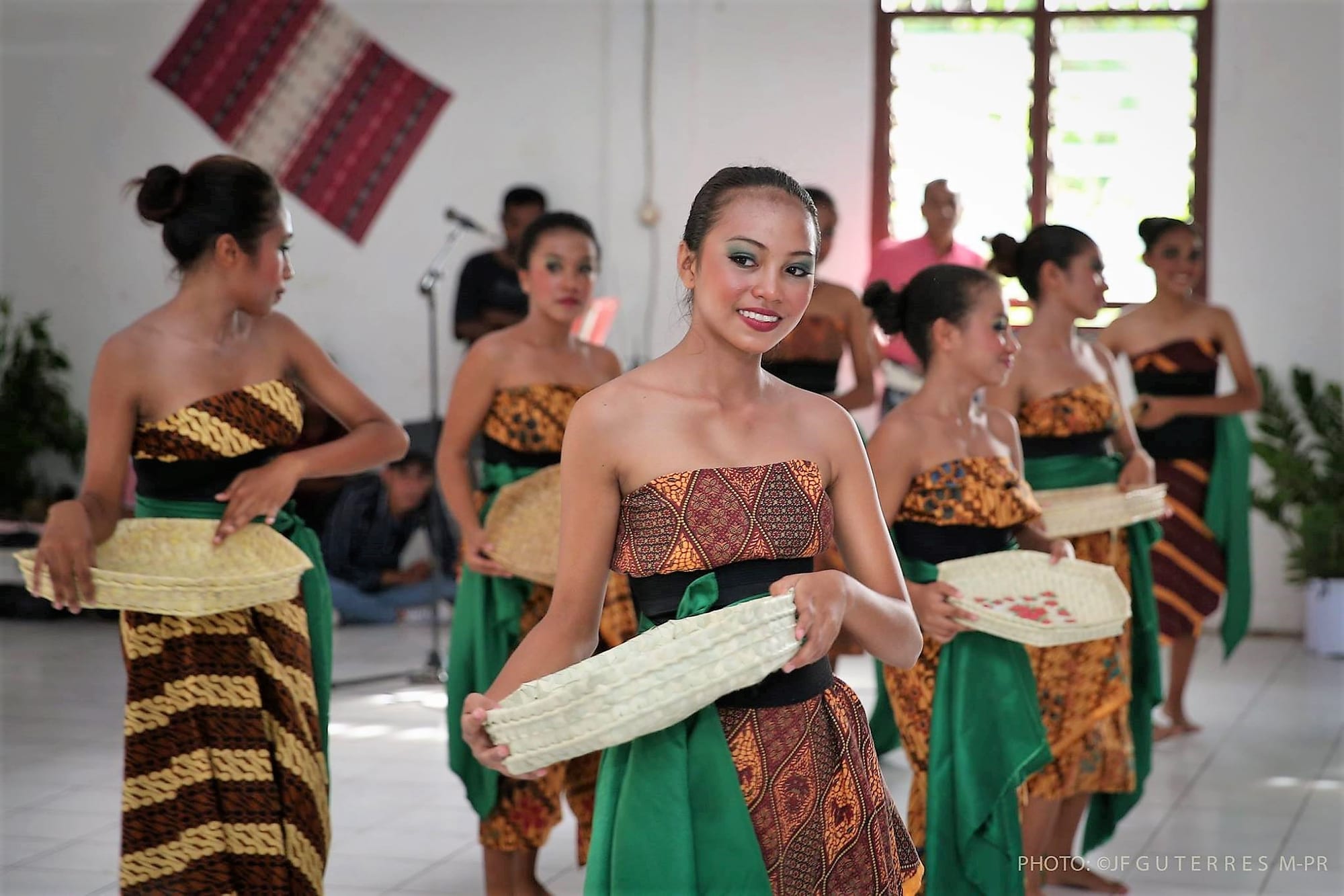 A row of young women in strapless patterned dresses with green sashes, each holding a shallow woven tray, performing a dance in a hall with a tais hanging on the wall behind them