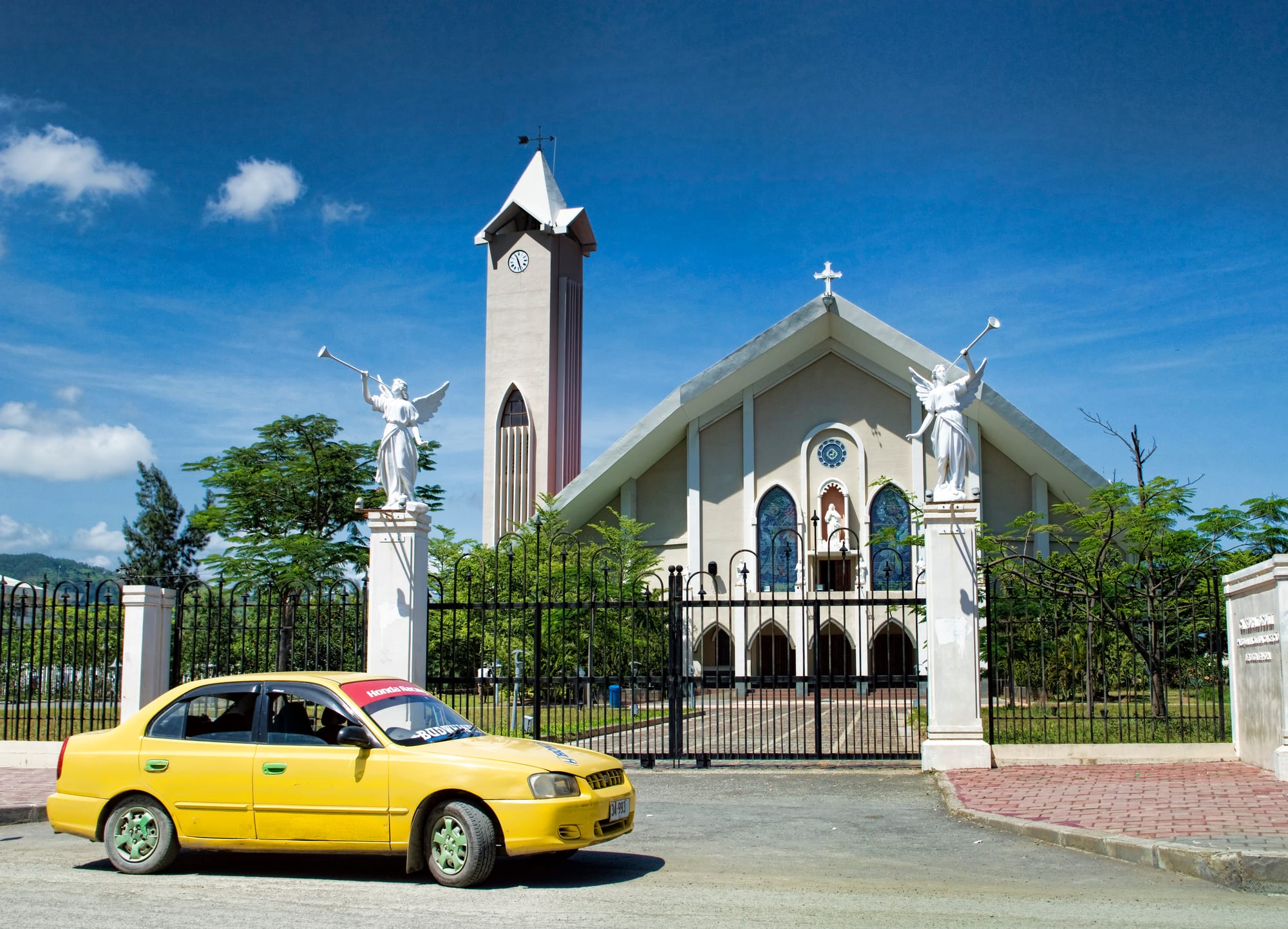 A pale cathedral with a steep gabled roof, blue stained-glass windows and a tall clock tower, seen through iron gates flanked by white angel statues, with a yellow taxi parked in front