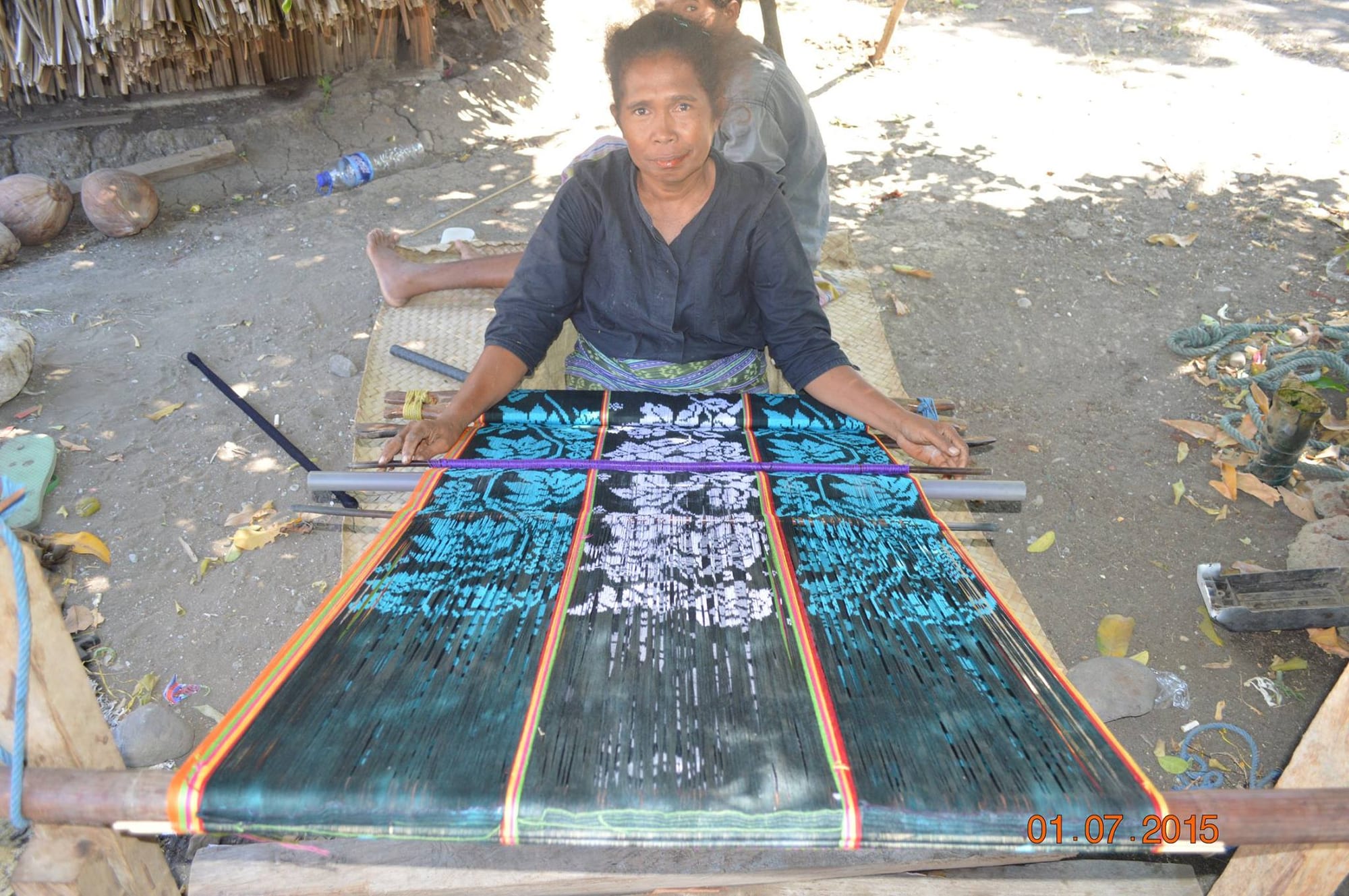 A woman sits on a mat on the ground behind a backstrap loom, weaving a dark tais with pale blue and white patterns and red and yellow edging