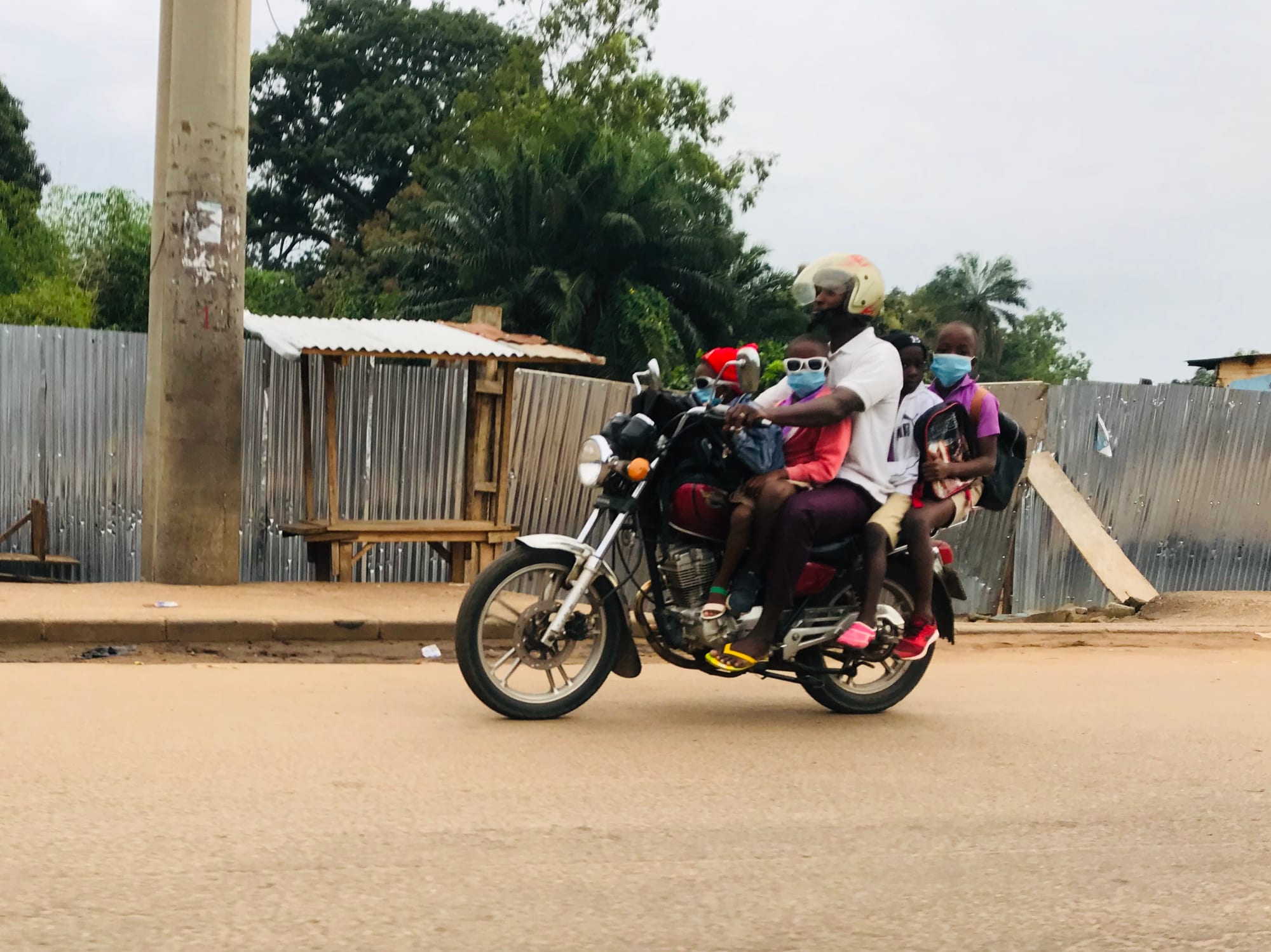 A man on a motorbike carrying four children in school clothes and backpacks along a dirt road